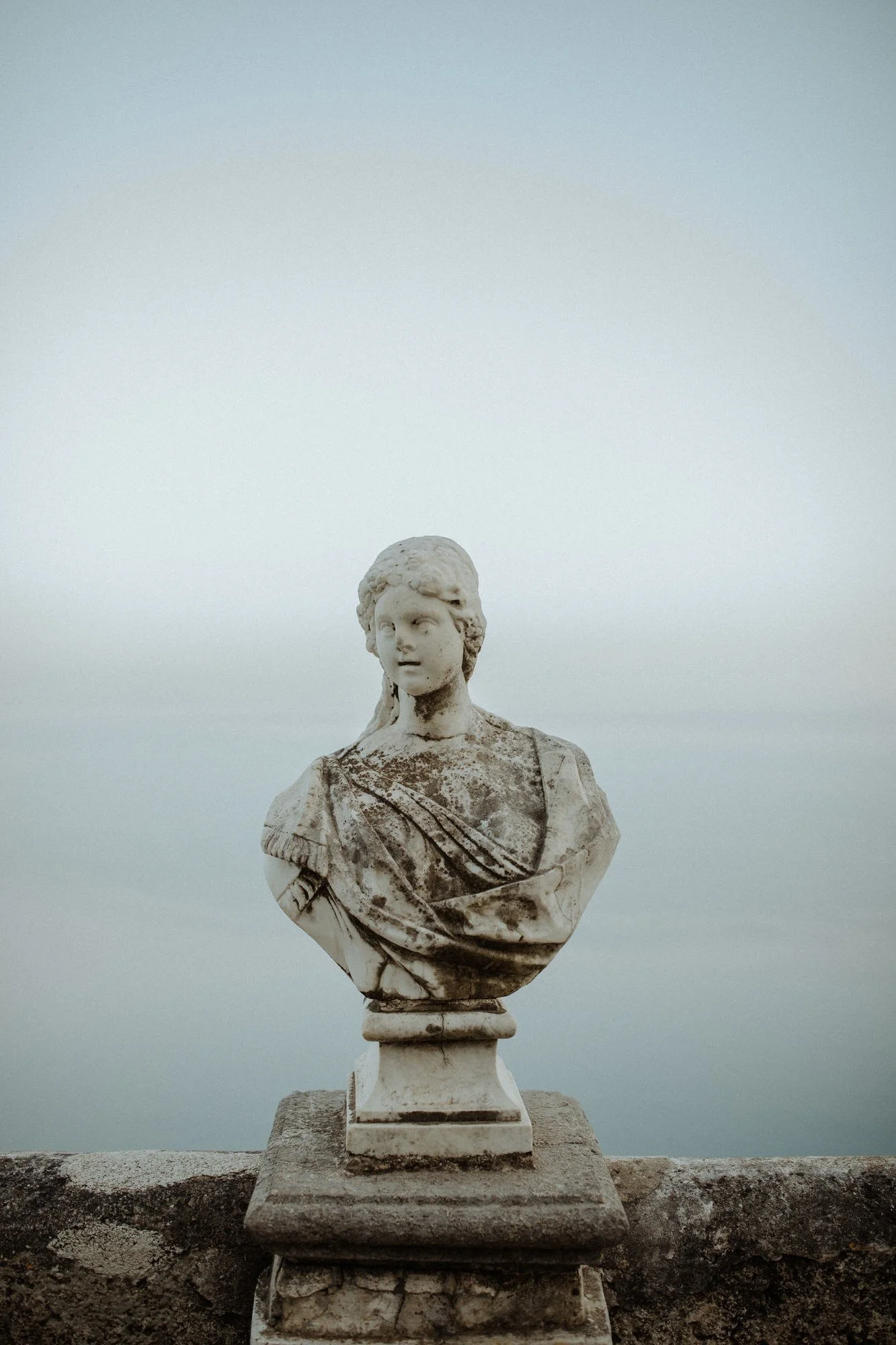 Weathered marble bust of a woman in draped robes, set on a stone balustrade against a pale sky at Villa Cimbrone, Italy.