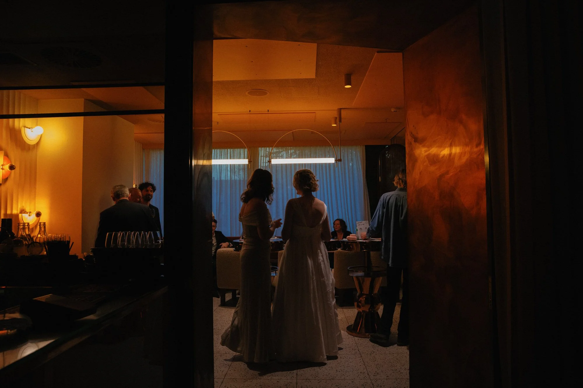 A bride and guest stand together in an intimate, warmly lit bar space, their dresses glowing softly in the amber light as guests gather around.