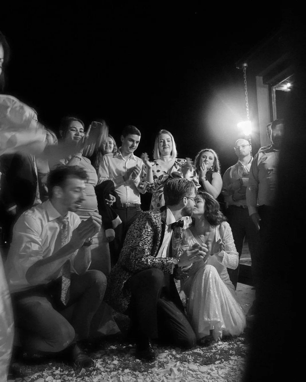 Petrichor Farm wedding reception in Canberra — a black and white candid of the bride and groom stealing a kiss while guests dance, clap, and cheer around them.