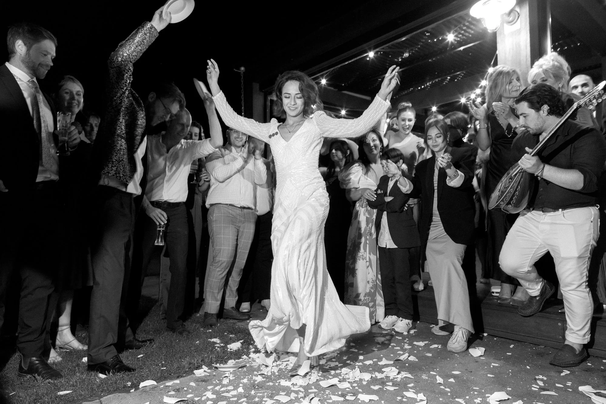 A bride dances through shards of broken plates as a circle of guests cheer and a musician strums — a wild, joyous nod to tradition and rebellion all at once.