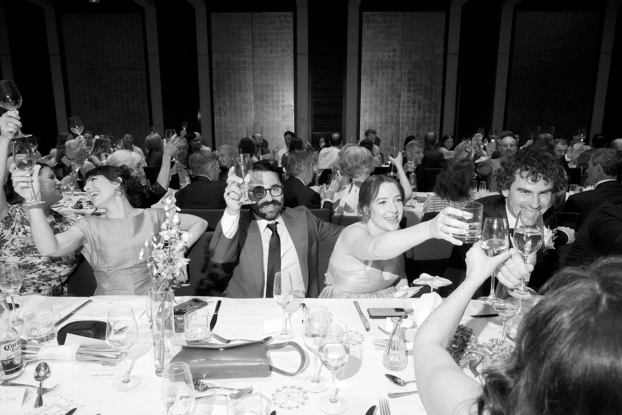 Wedding guests raising glasses in a joyful toast during a reception at Gandel Hall, National Gallery of Australia in Canberra, captured in black and white by Corinna & Dylan, modern wedding photographers.