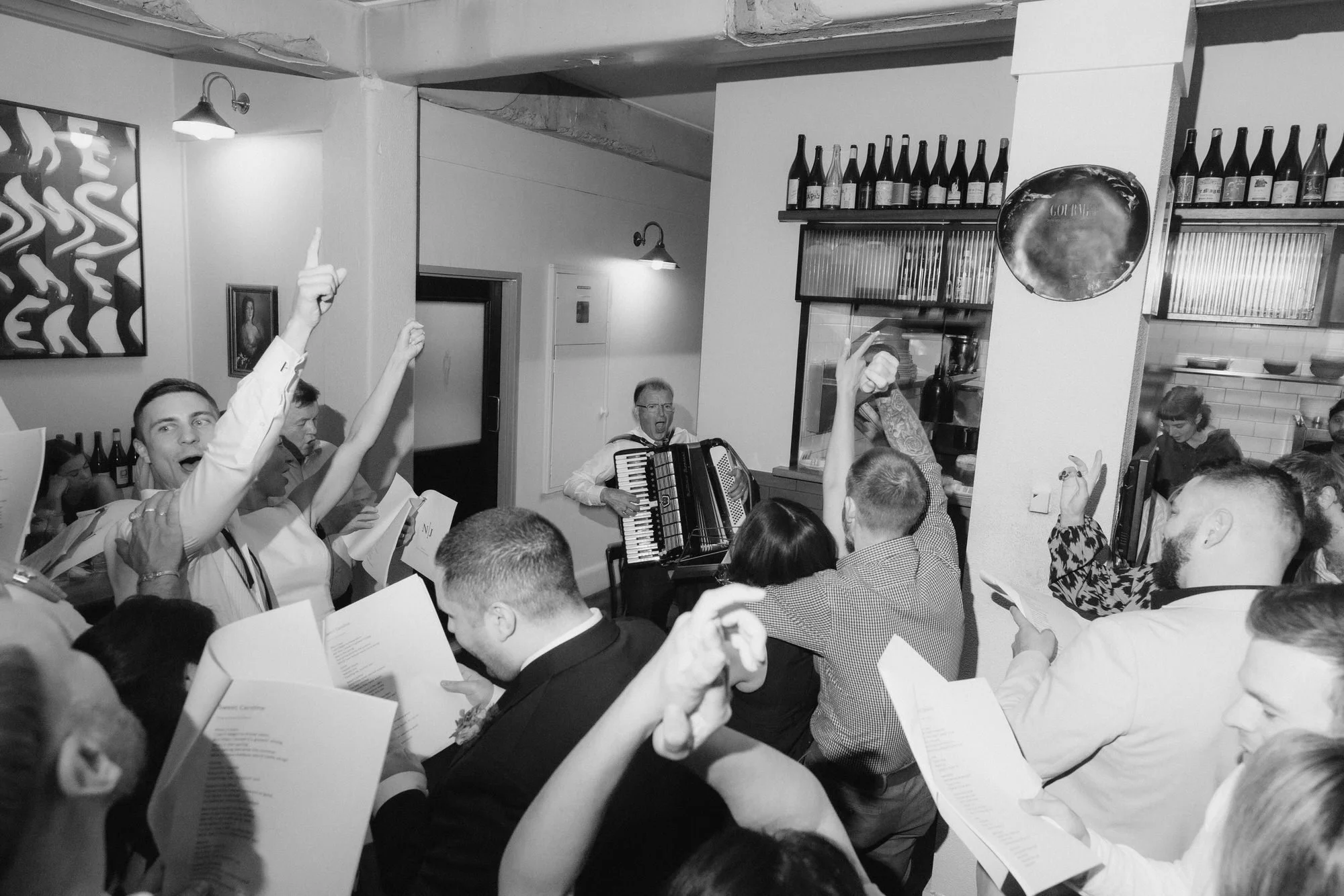 Wedding guests singing loudly with lyric sheets as an accordion player leads the party at Bar Rochford, Canberra — a lively, joy-filled moment of community and music
