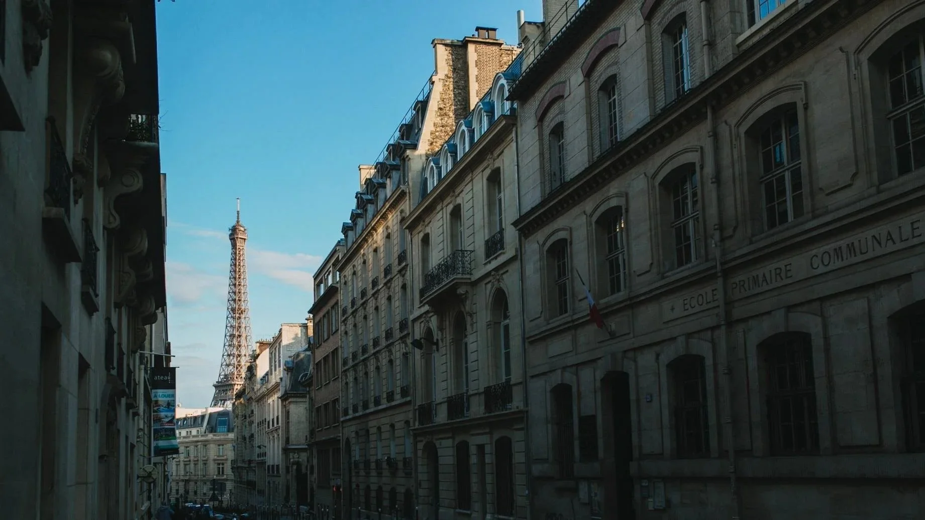 A quiet Parisian street with the Eiffel Tower rising in the distance, framed by rows of classic stone buildings and a clear evening sky