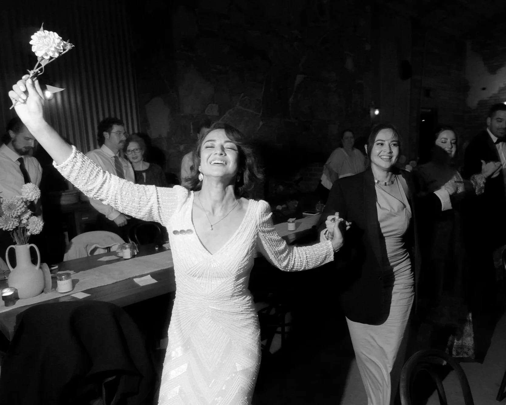 A bride, eyes closed and face lifted with joy, dances with guests during a lively wedding celebration, holding a flower aloft