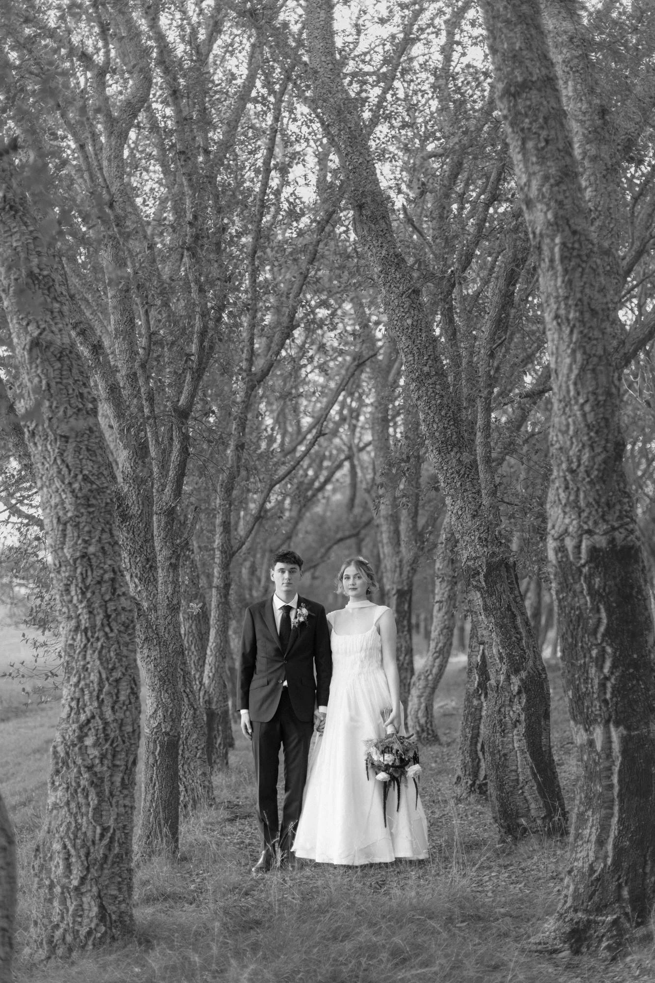 Bride and groom stand together in a grove of tall trees, holding hands. The black-and-white edit enhances the timeless, cinematic feel, with soft tones that emphasize the textures of the trees and the flowing gown