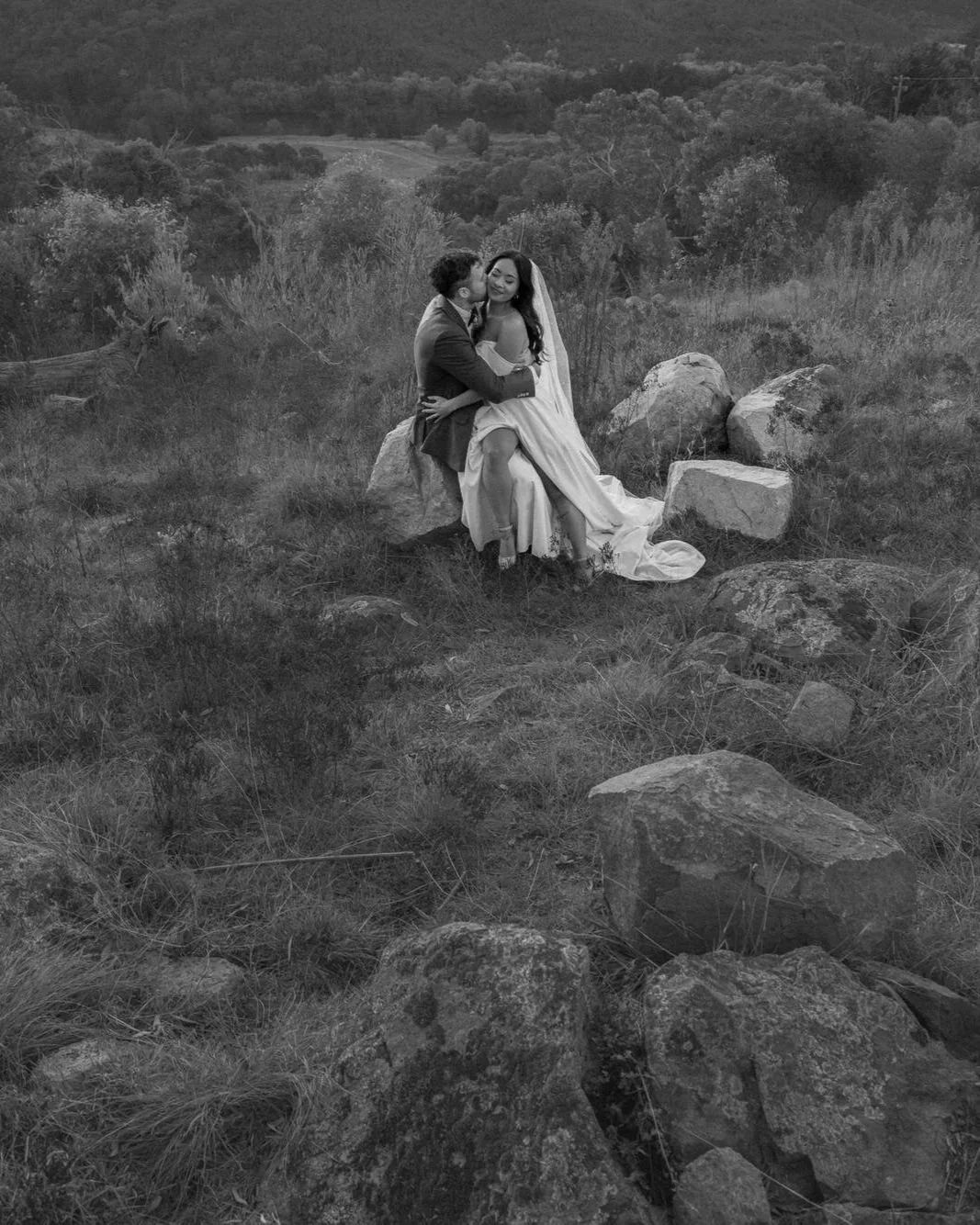 Two newlyweds folded into each other among the boulders of Brindabella Hills — the dress spilling over stone, the bush rising around them, and the mountains holding them in quiet witness.