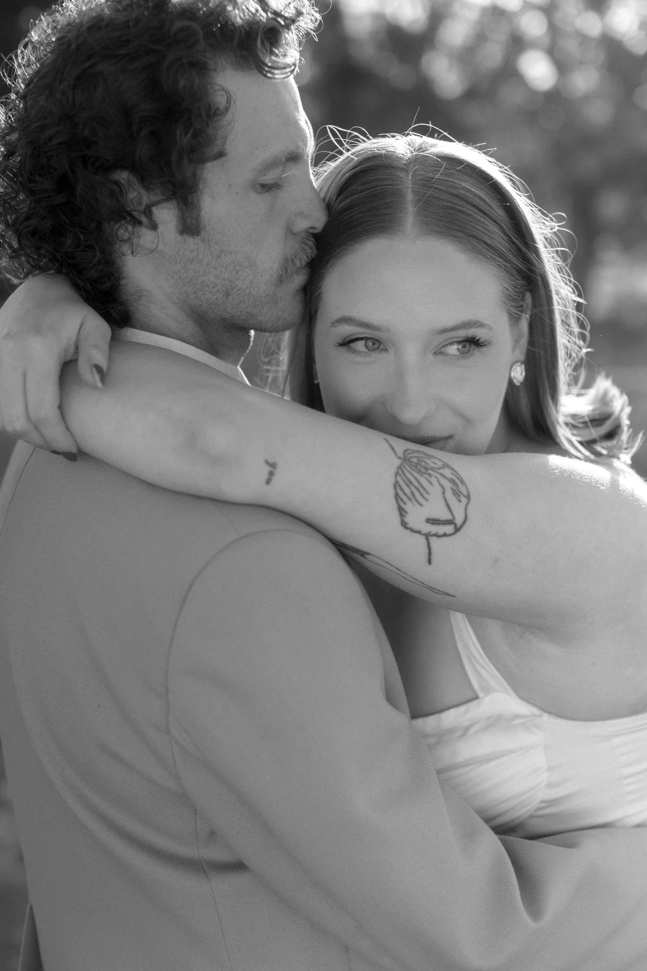 A quiet, intimate wedding portrait — her arm draped around his shoulder, tattoo catching the light, as he presses a kiss to her temple.