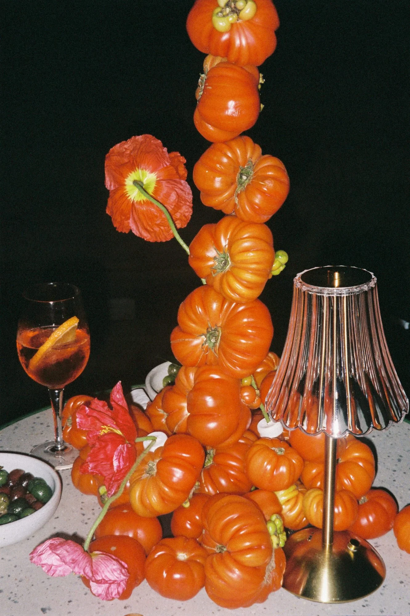 A wedding centerpiece made of stacked tomatoes and bold red flowers, styled on a table with cocktails, olives, and candlelight. 35mm film.