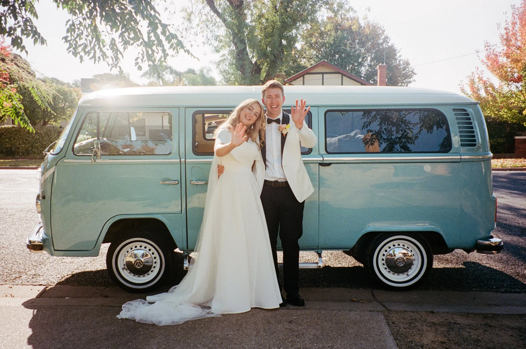 Newlyweds waving cheerfully while standing in front of a vintage pale blue kombi van, captured on 35mm film