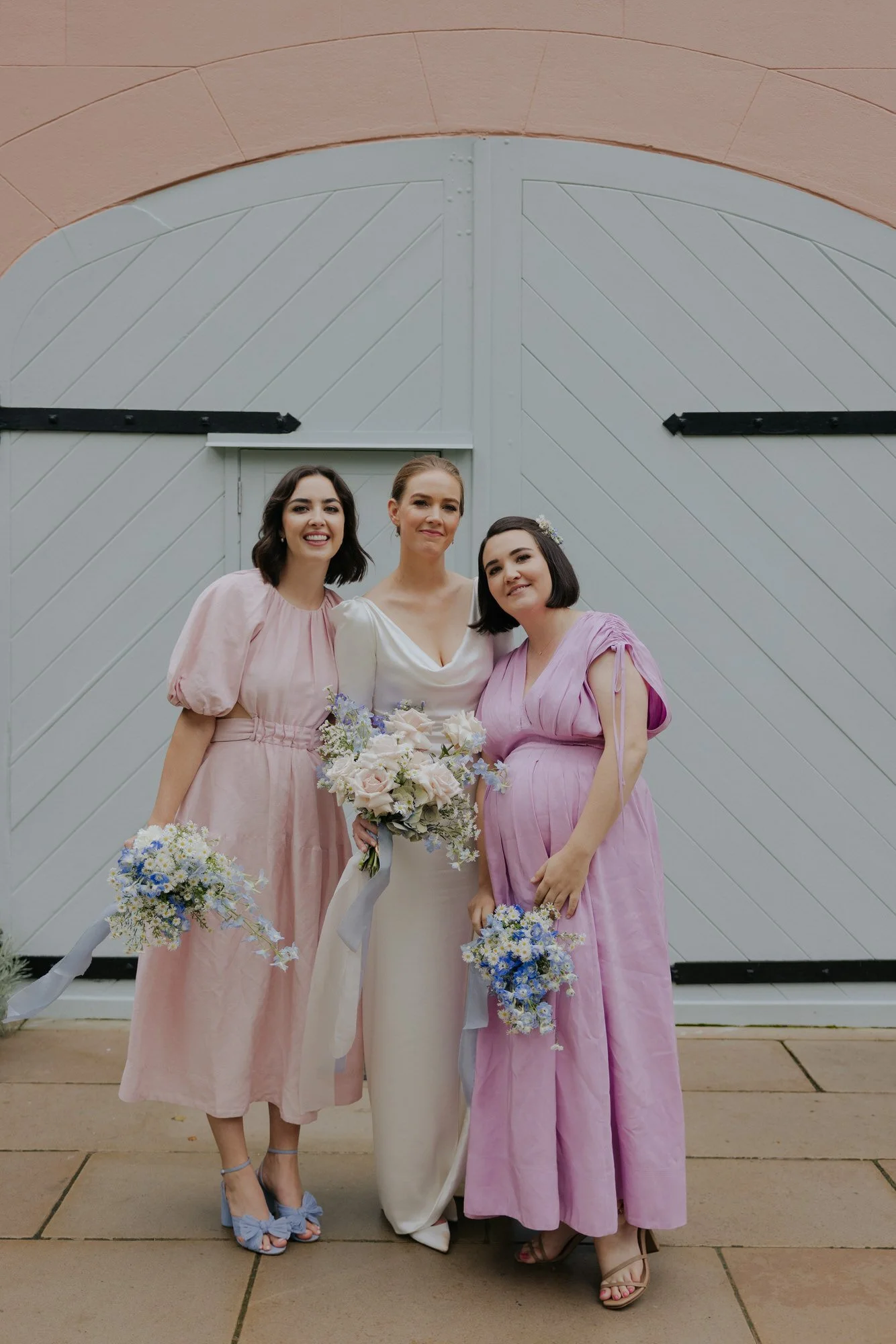 Bride standing with two bridesmaids in pastel pink dresses, each holding delicate bouquets of soft pink, blue, and white flowers, against the pale timber barn doors at Mona Farm