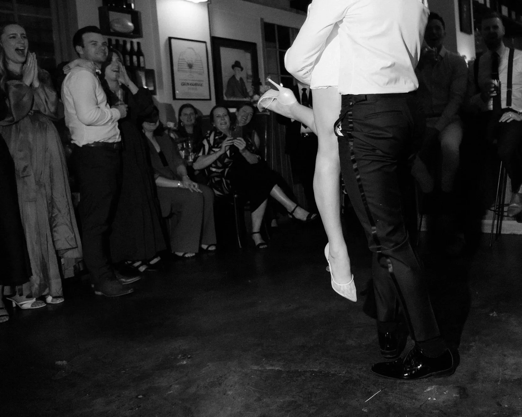 The floor of Bar Rochford becomes a stage — her heel lifted mid-air, their embrace pulling cheers from friends pressed close in the dim-lit room. A first dance that feels more like a secret shared than a performance.