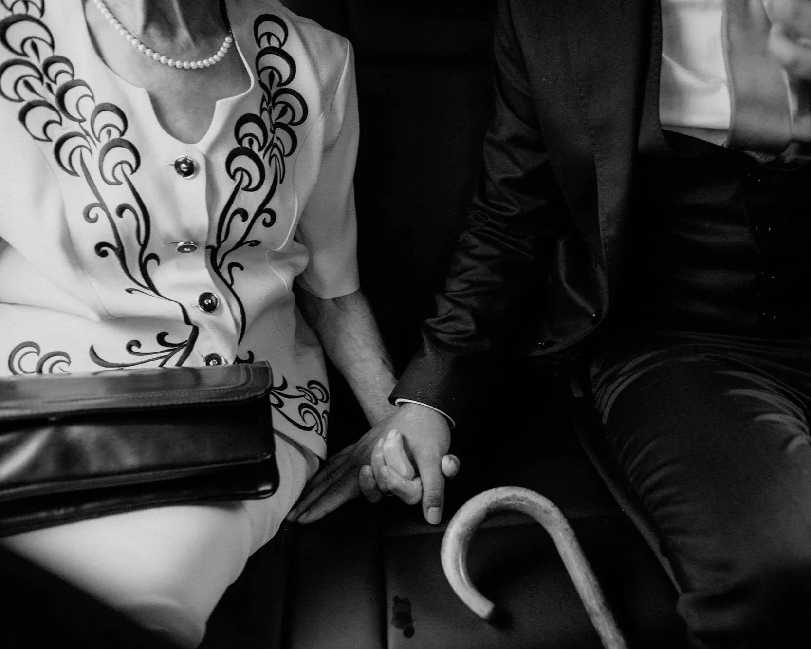 A tender close-up of the groom and his grandmother holding hands in the back of a car, her cane resting nearby, capturing a moment of quiet connection on the wedding day.