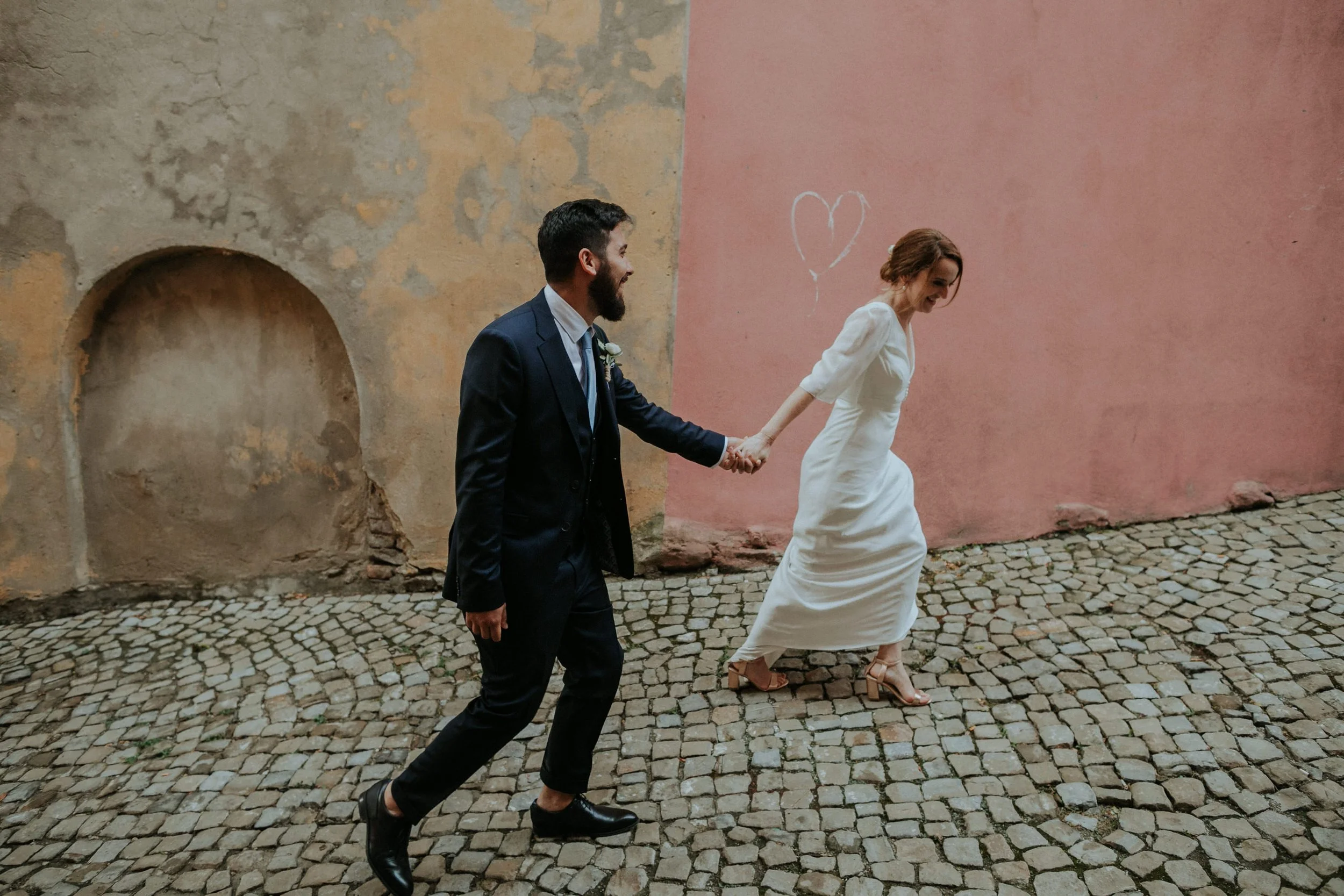 Playful destination wedding photo in Monforte d’Alba, Italy — bride in a simple white dress leads her groom along a cobblestone street with a pink wall and heart graffiti in the background