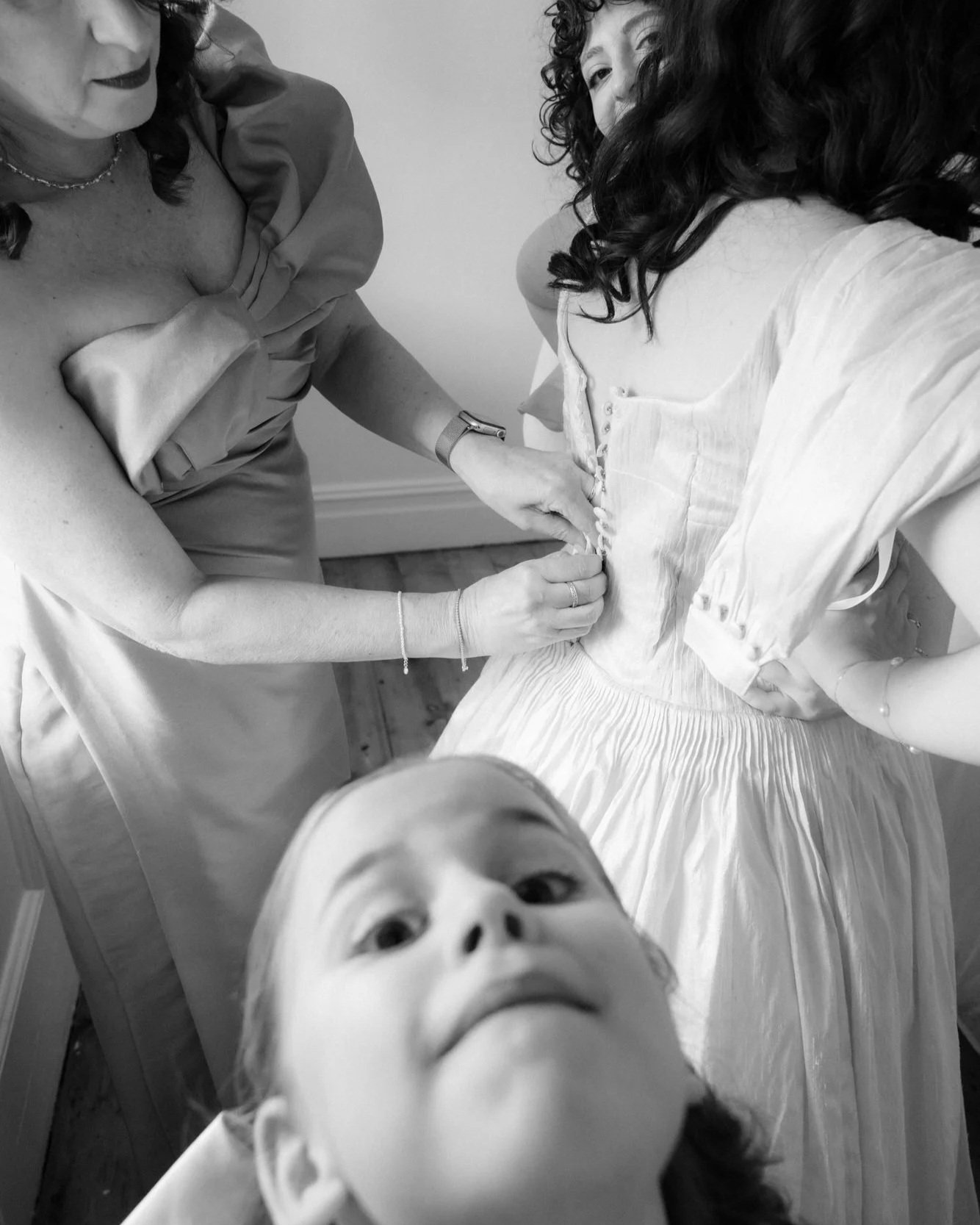 A dress being buttoned, hands working delicately, while a child pops into frame with wide-eyed mischief — the poetry of wedding prep colliding with play.