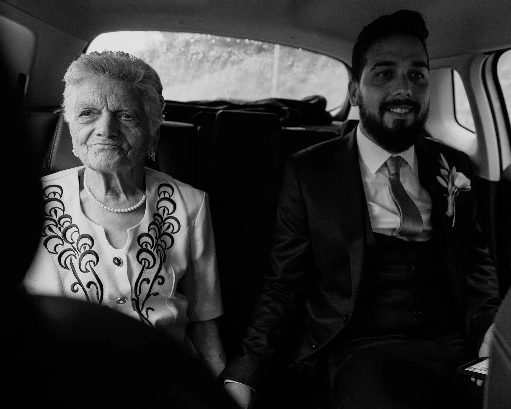 The groom sits beside his grandmother in the back seat of a car, dressed for the wedding. She wears pearls and a patterned jacket, their presence quiet but full of meaning.