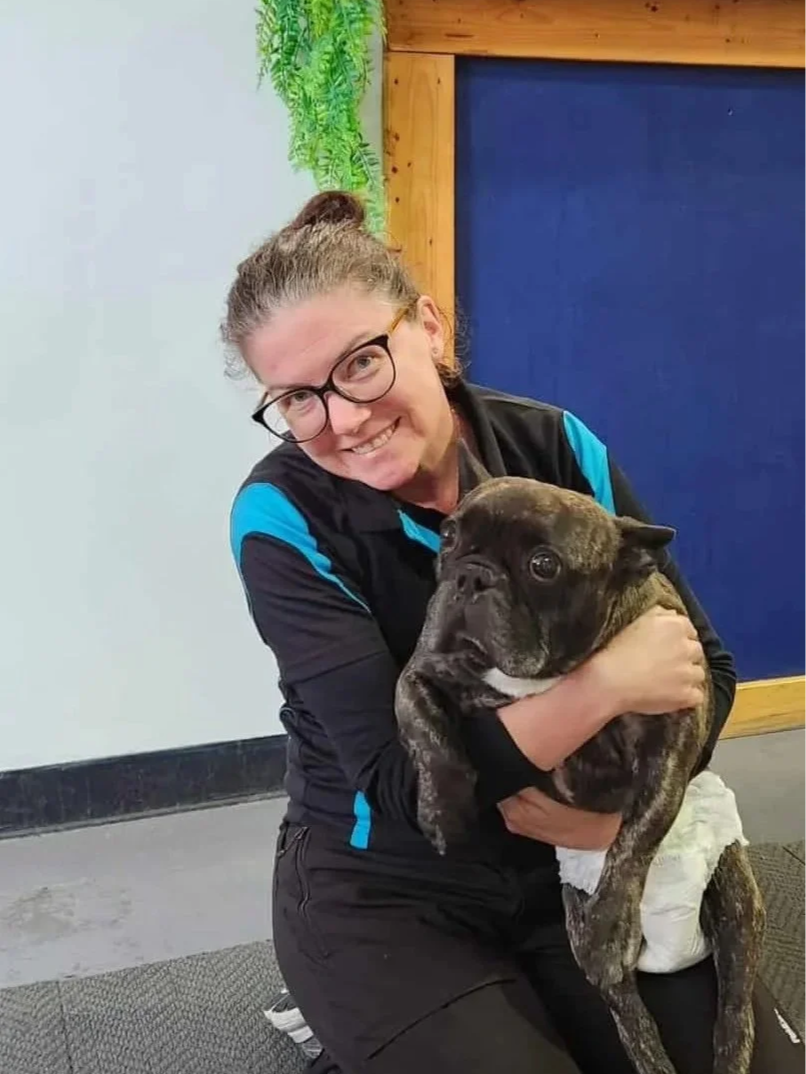 Alicia smiling while holding a brindle French Bulldog puppy indoors at Water Walkies Port Kembla against a wall with green plants and wood paneling.