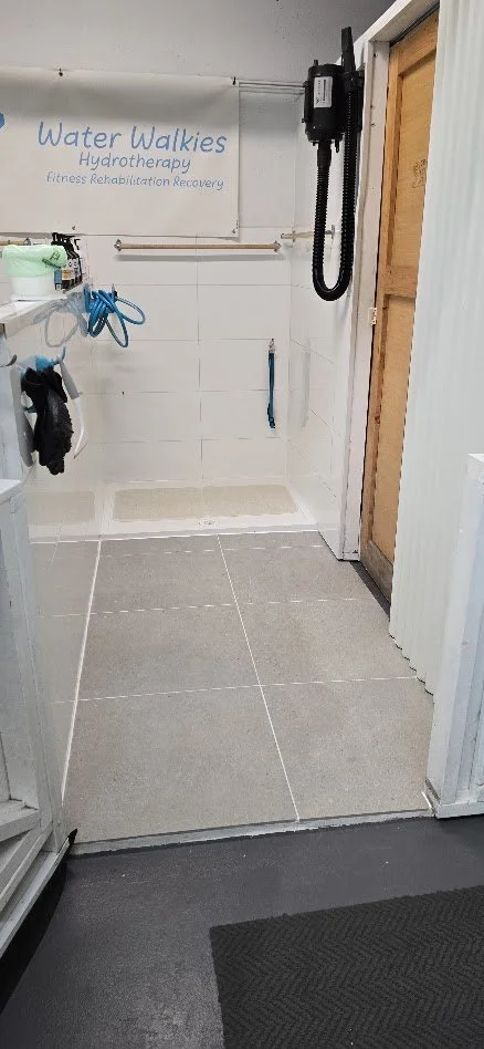 The DIY dog wash bay designed for water walkies hydrotherapy, with a small tiled area, water equipment on the walls, and a sign that reads 'Water Walkies Hydrotherapy Fitness Rehabilitation Recovery.'