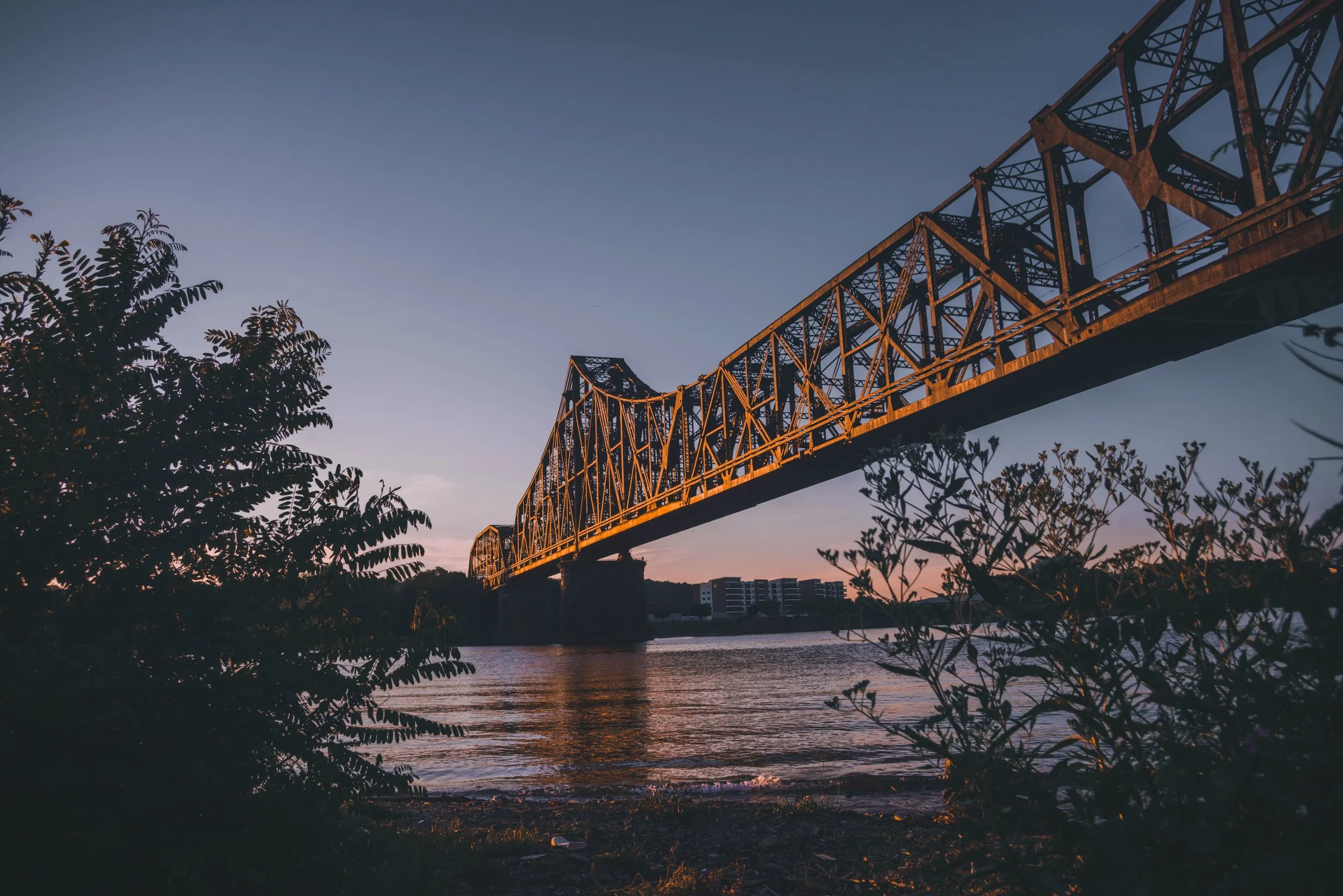 A large, steel truss bridge spans over a body of water at sunset, with trees and buildings visible below and in the background.