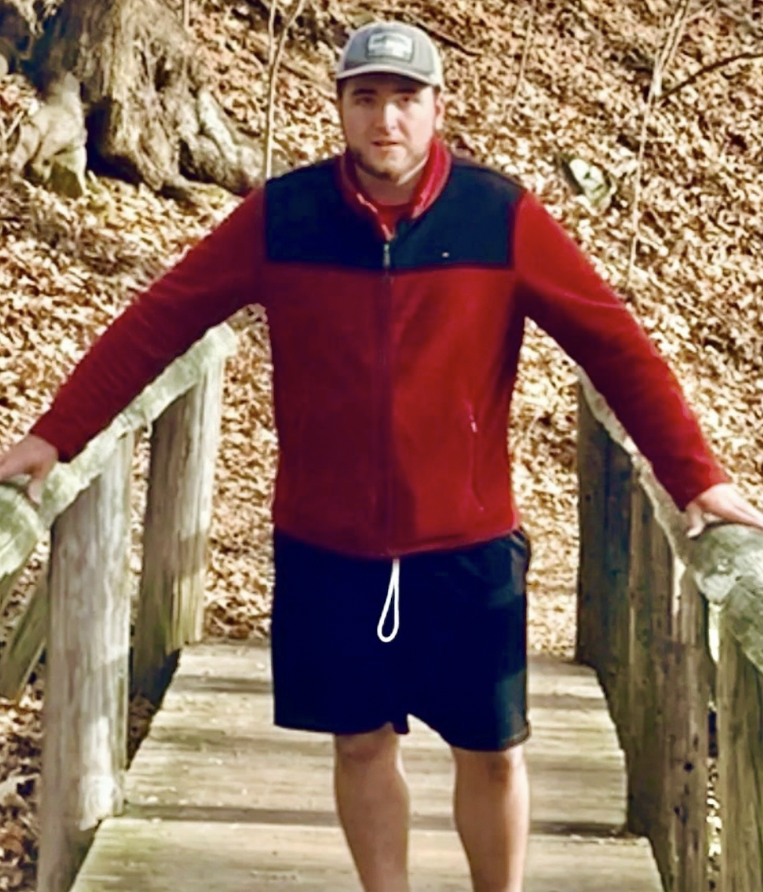John Ruscik of Greenville wearing shorts, a hat, and a red jacket on a hike standing in the middle of a bridge.