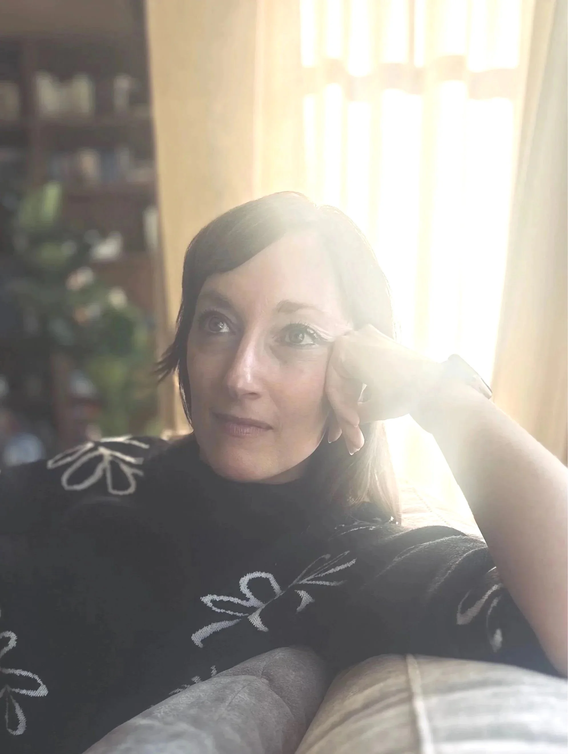A woman with dark hair and blue eyes with her head resting on her hand, looking off to the side, in a room with sunlight streaming through curtains behind her. Online therapy for moms, parents and people in perimenopause in California.