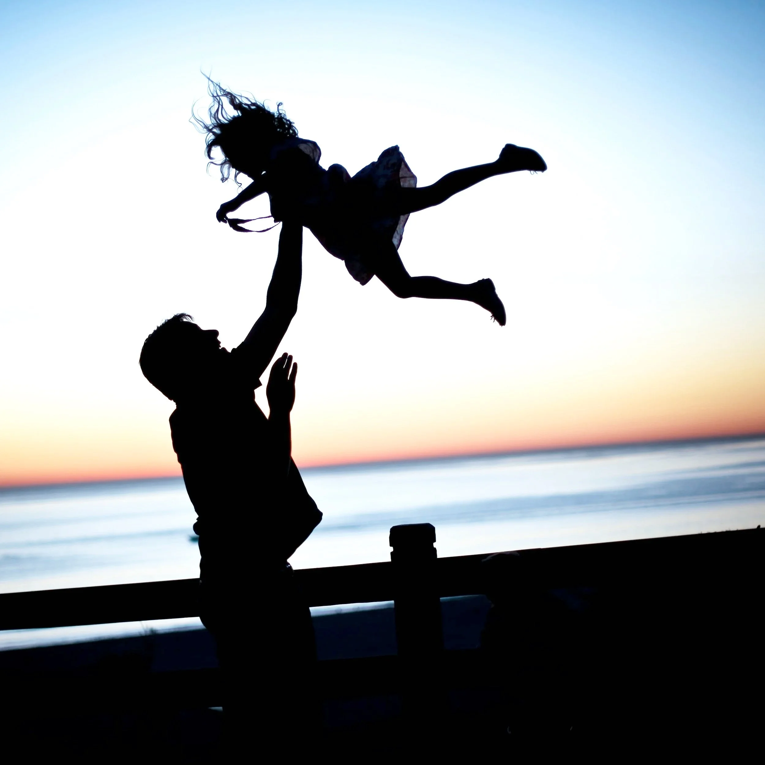 Silhouette of a man tossing a young girl into the air at sunset by the beach. Postpartum depression therapy online in California.