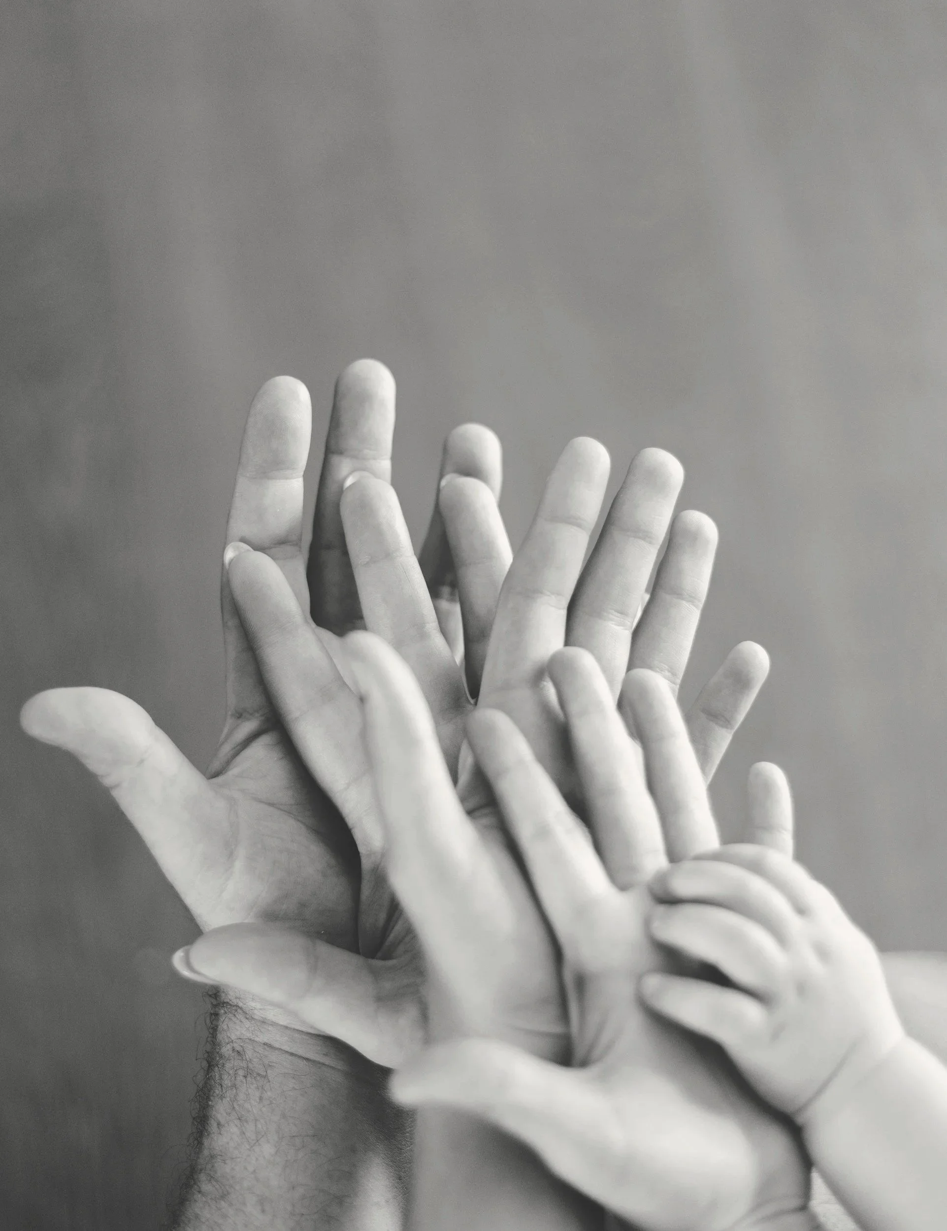 Group of multiple human hands of different sizes, including a small child's hand, stacked together against a plain background.