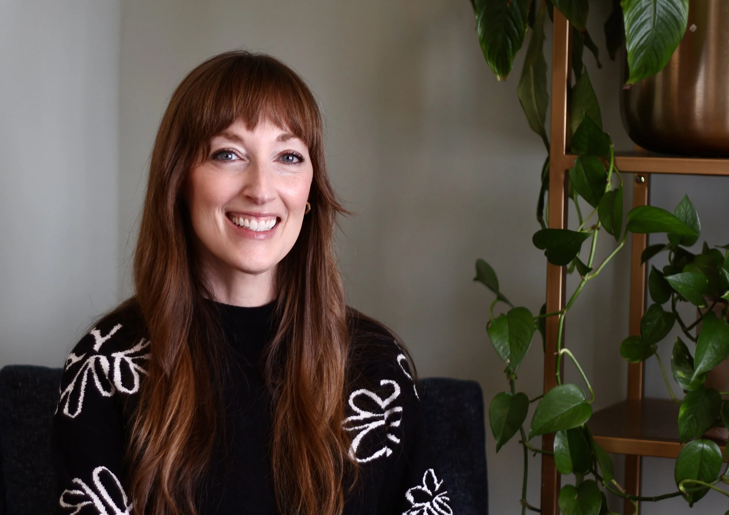 A woman with long, wavy red hair and a black sweater with white floral patterns, smiling while sitting indoors near a plant in a room with green walls. Online therapy for postpartum anxiety and depression in California.