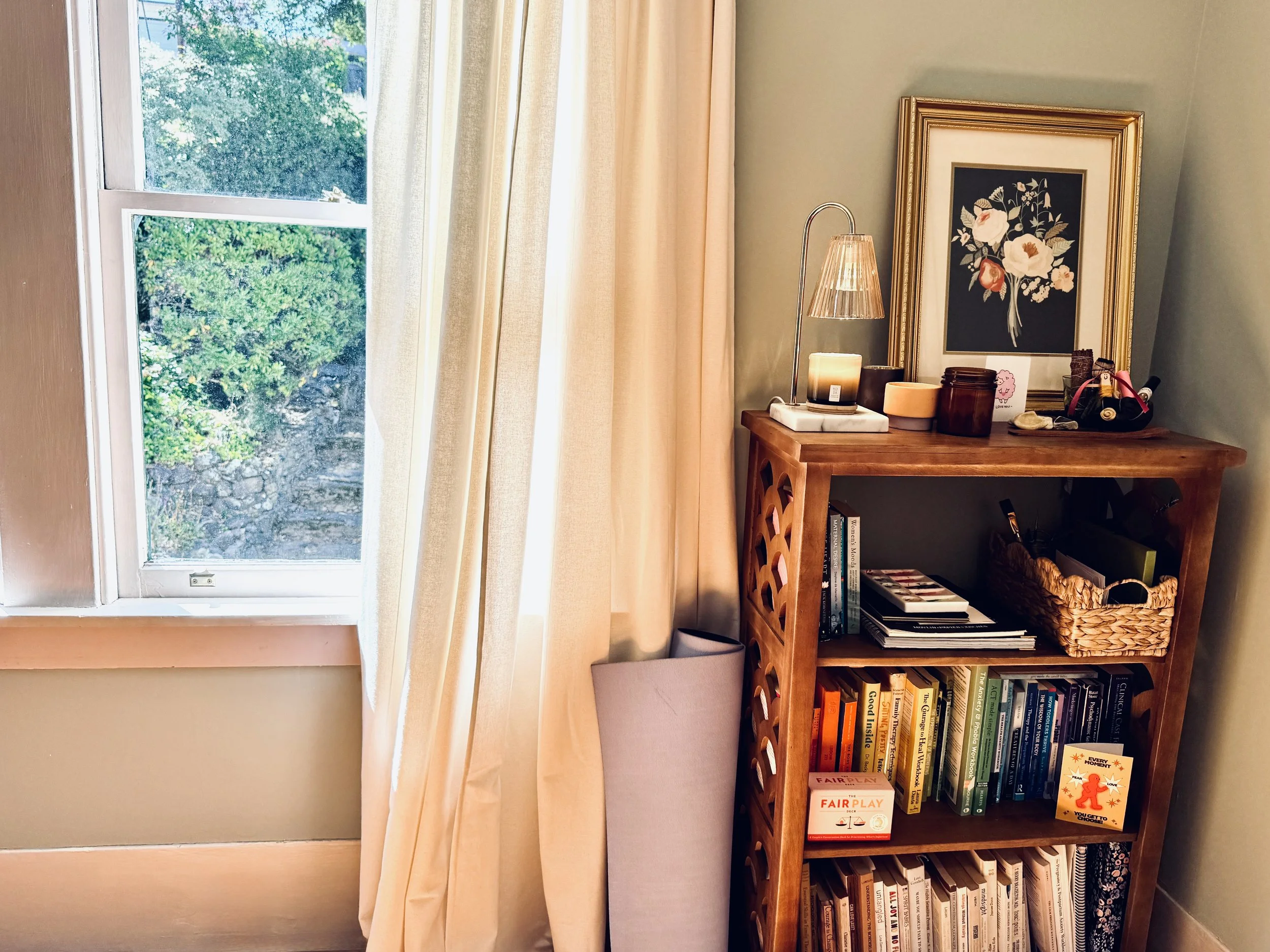 A cozy corner of a room with a bookshelf filled with books, a framed floral art print hanging, a small table lamp, candles, and decorative objects, next to a window with a view of green foliage outside. Therapy for new parents in California.