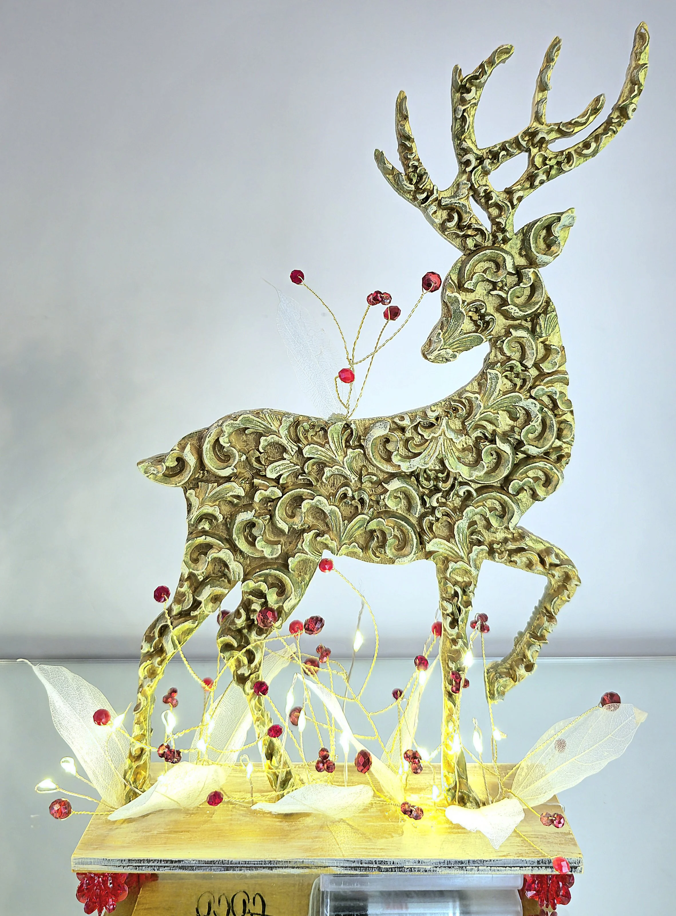 Statue de renne en or ornée de décorations rouges et blanches, avec des lumières intégrées, sur une base en bois.