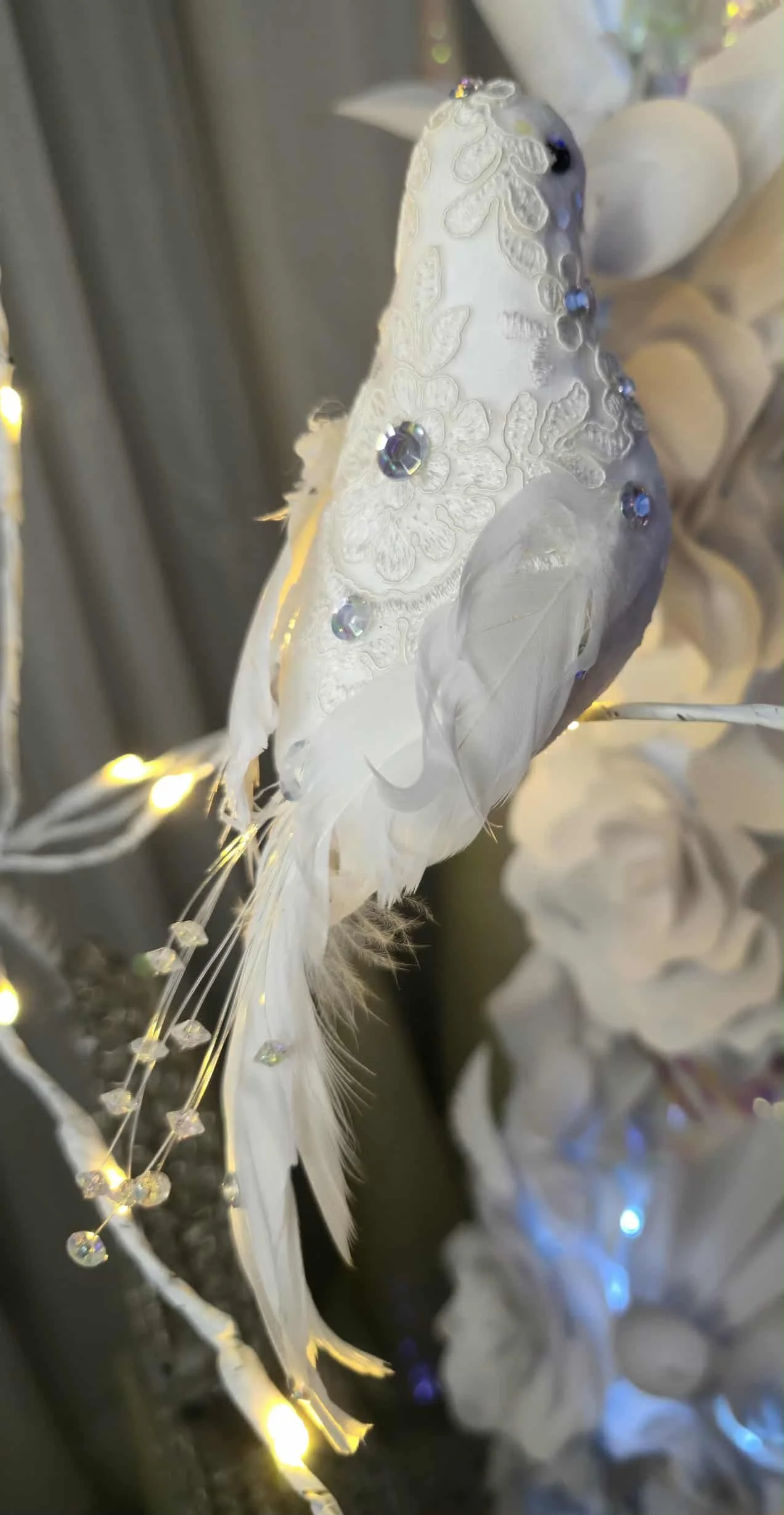 Un oiseau décoratif blanc avec des plumes, orné de cristaux et éclairé par des petites lumières LED, suspendu parmi des branches blanches.