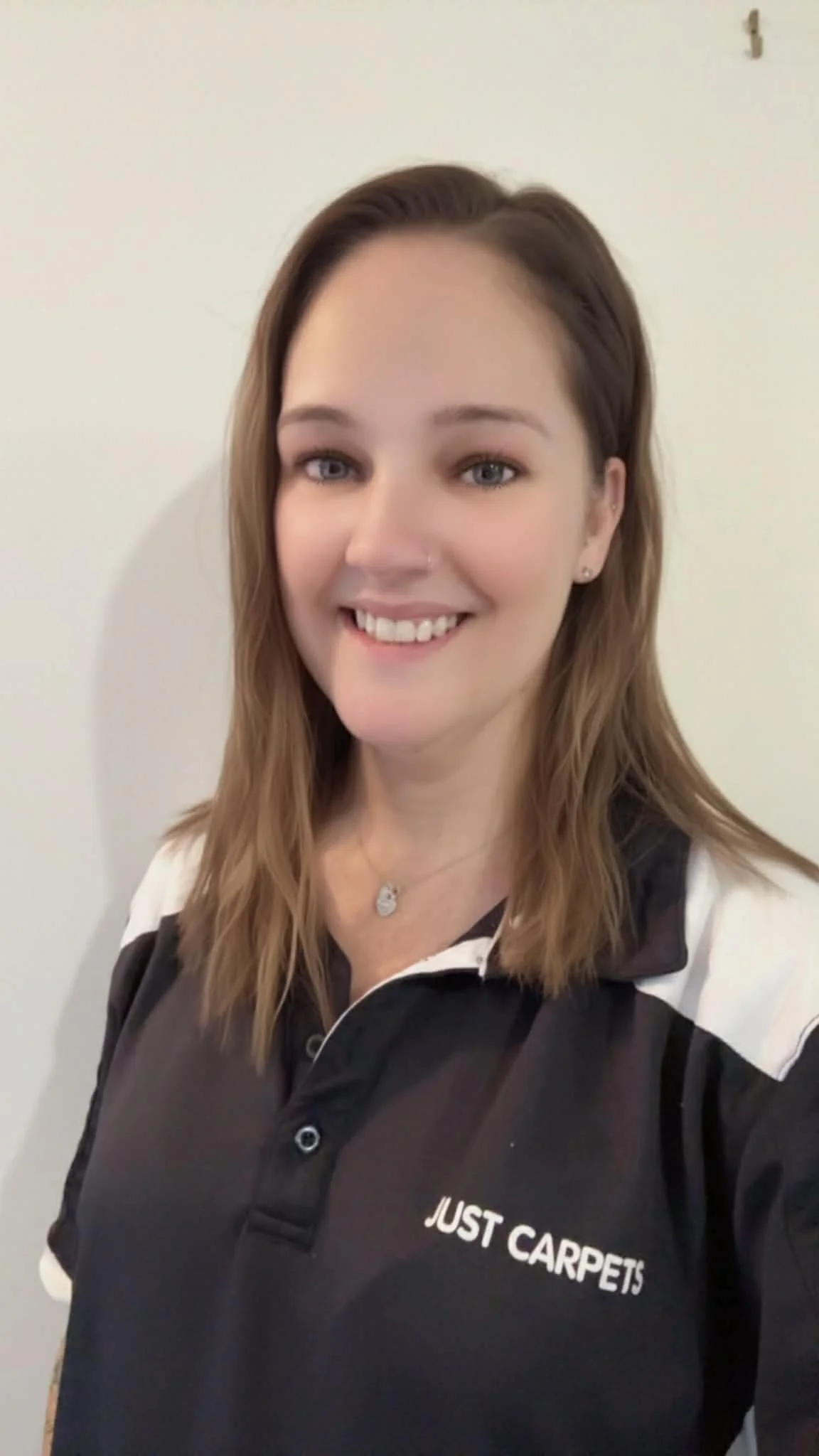 A woman smiling wearing a black and white shirt with a logo that says 'JUST CARPETS', standing against a plain white wall.