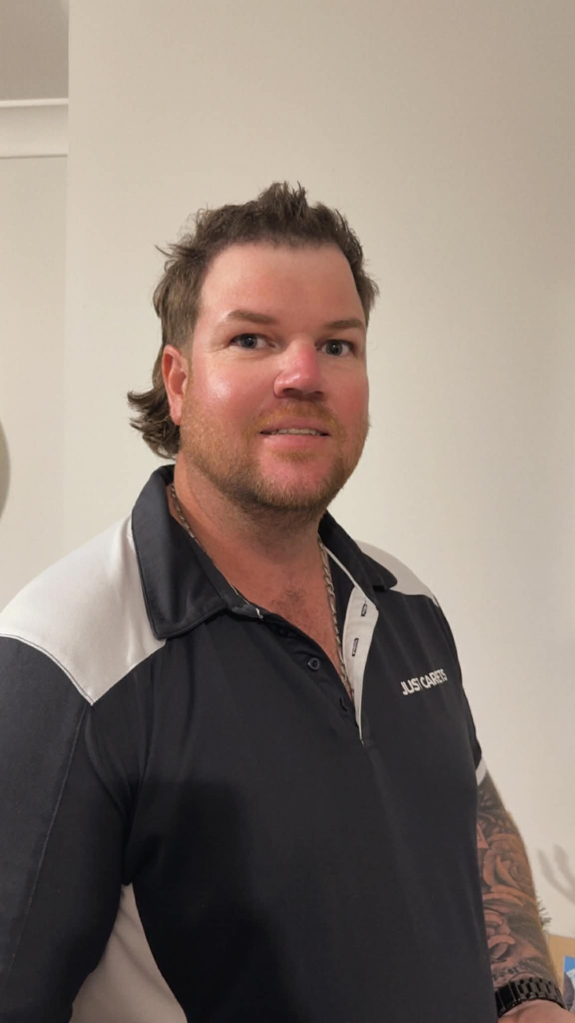 A man with medium-length brown hair, blue eyes, and a beard, wearing a black and white polo shirt with the words 'JUST CARES' on it, standing in front of a plain wall.
