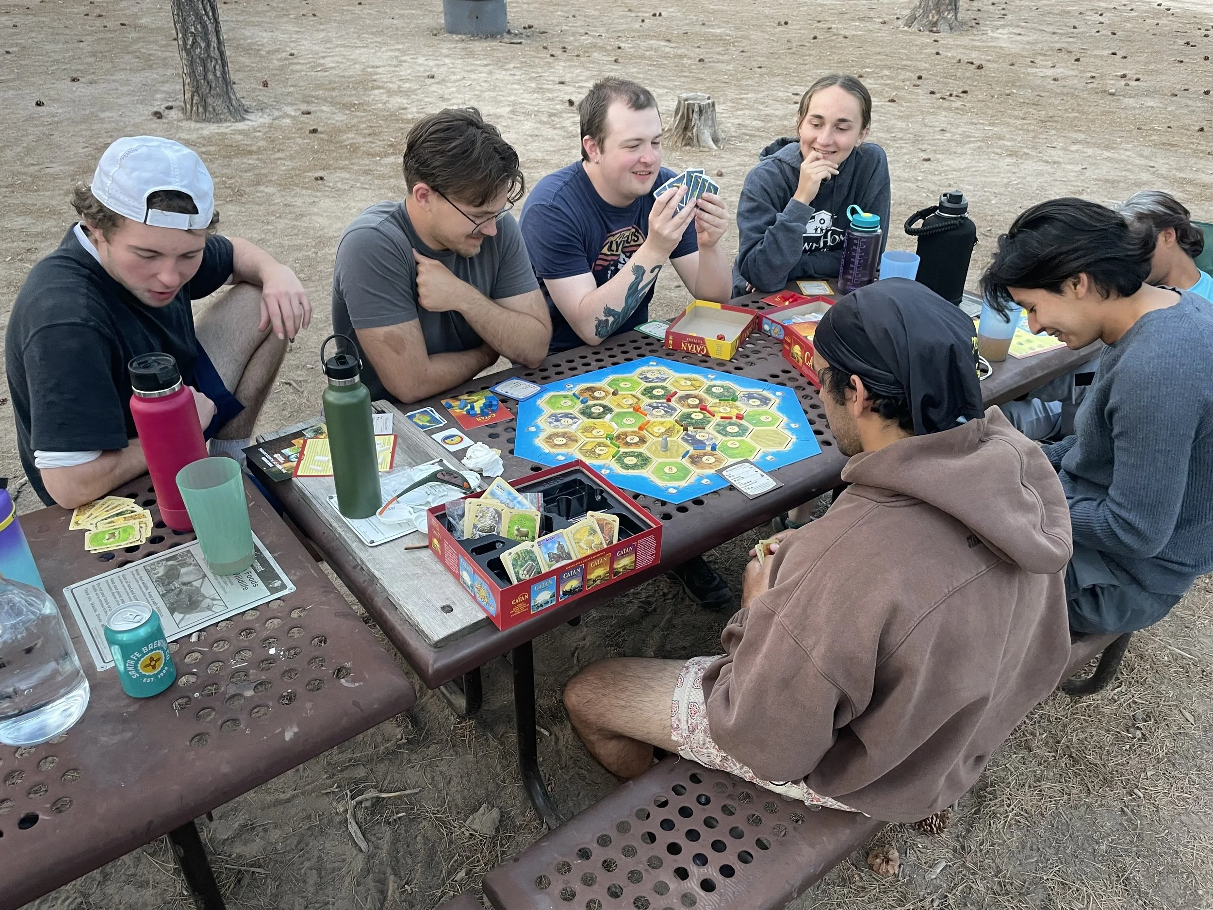 SAGE, Summer of Applied Geophysical Experience. Student group playing the board game "Catan" outdoors at a picnic table surrounded by trees.