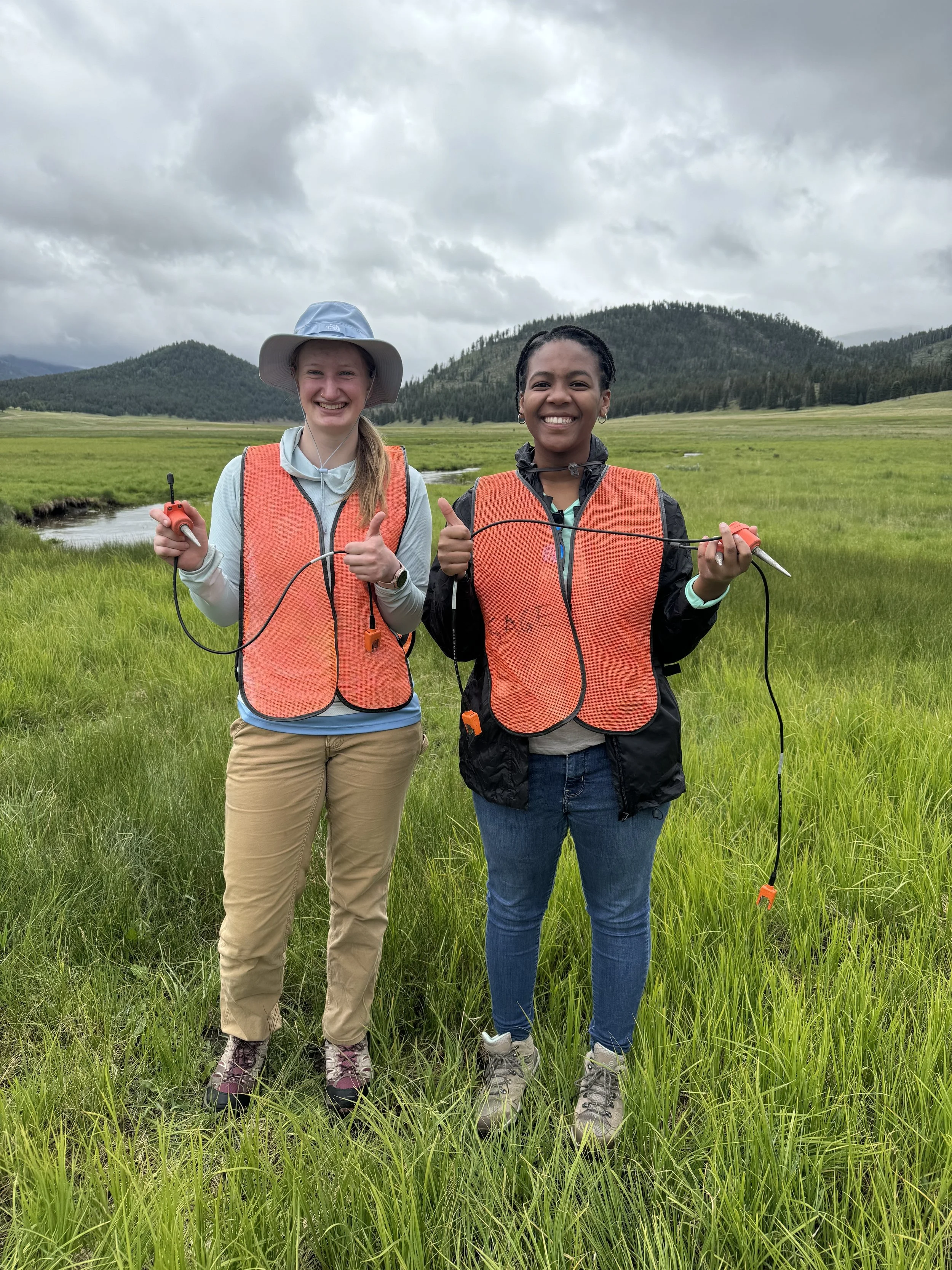 SAGE, Summer of Applied Geophysical Experience. Two women in outdoor field wearing orange safety vests holding environmental sampling equipment.