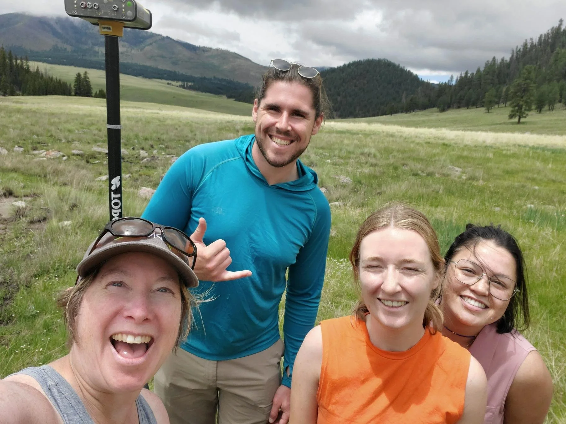 SAGE, Summer of Applied Geophysical Experience. Four smiling people outdoors in a grassy field with mountains and trees in the background. 