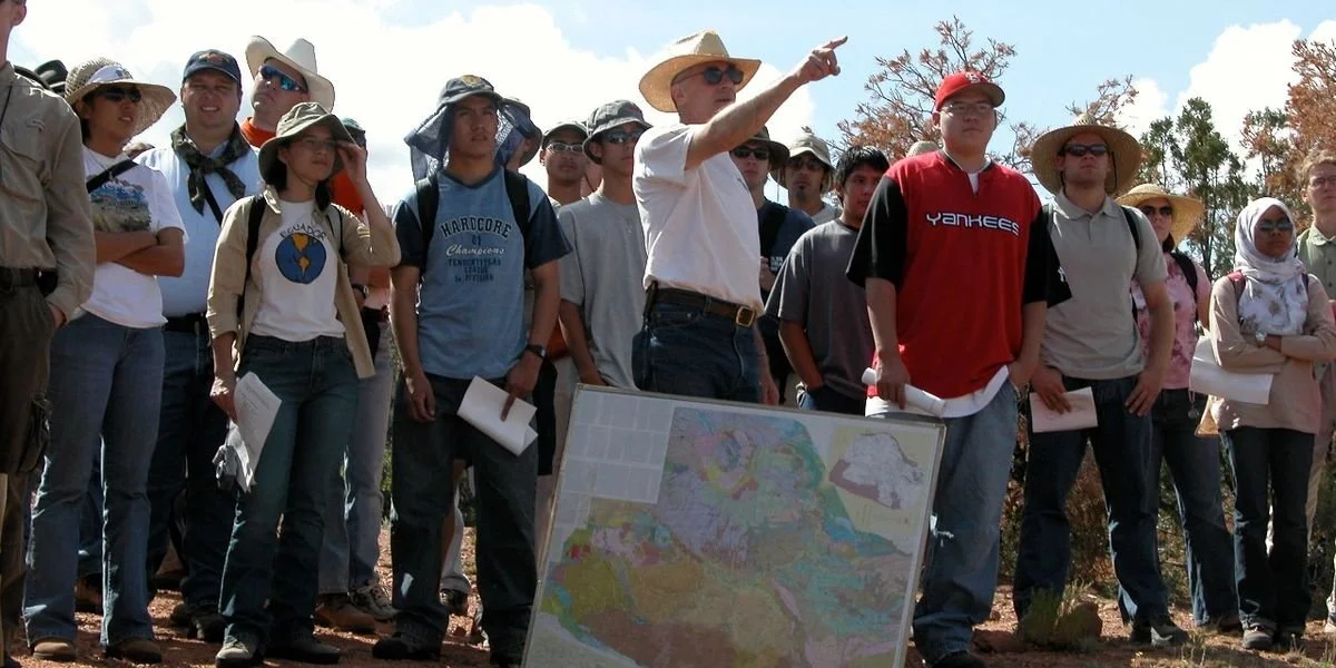 SAGE, Summer of Applied Geophysical Experience. Group of people outdoors, listening to an instructor pointing, with a map on a stand in front of them.