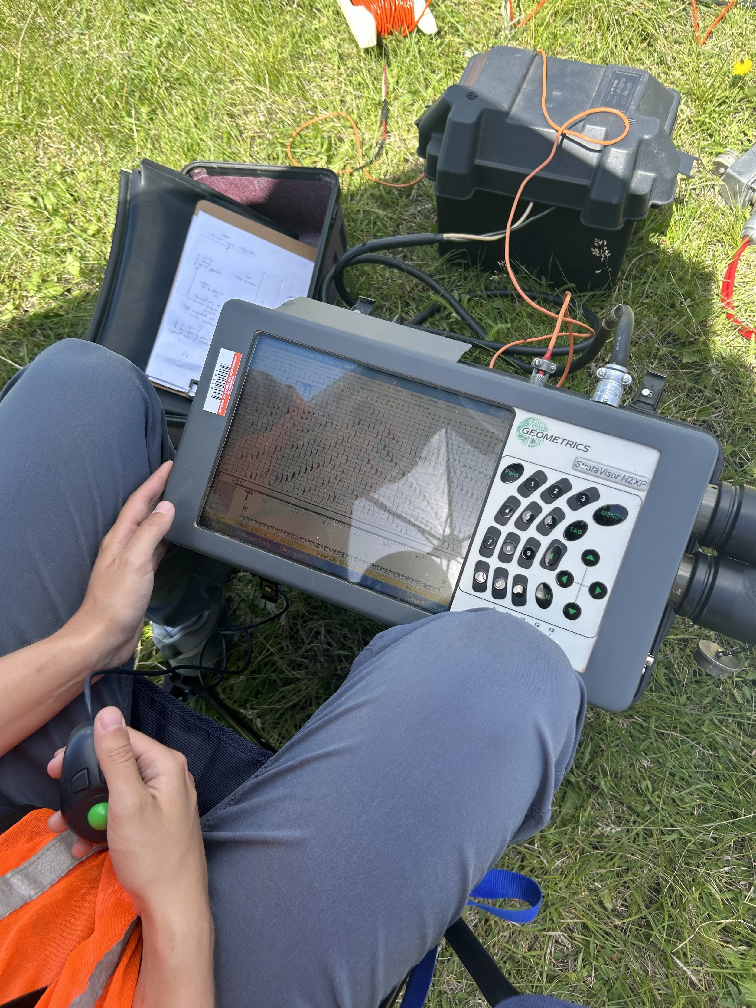 SAGE, Summer of Applied Geophysical Experience. Person holding a portable seismograph device outdoors on grassy ground. The device displays a seismic graph.