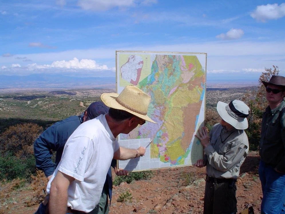 SAGE, Summer of Applied Geophysical Experience. Group of people outdoors in a hilly, semi-arid area, examining a large topographic map on an easel, with a view of distant hills under a partly cloudy sky.
