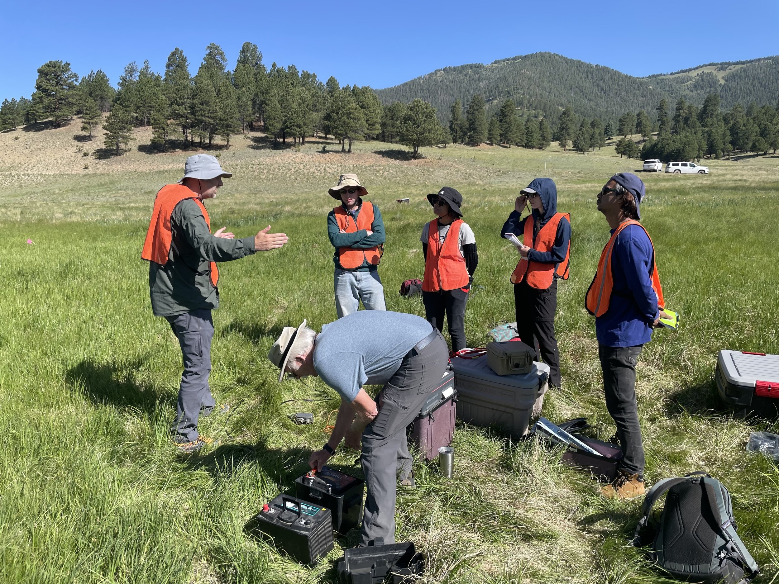 SAGE, Summer of Applied Geophysical Experience. Research team conducting fieldwork in a grassy meadow surrounded by trees and hills, with equipment and vehicles visible in the background.