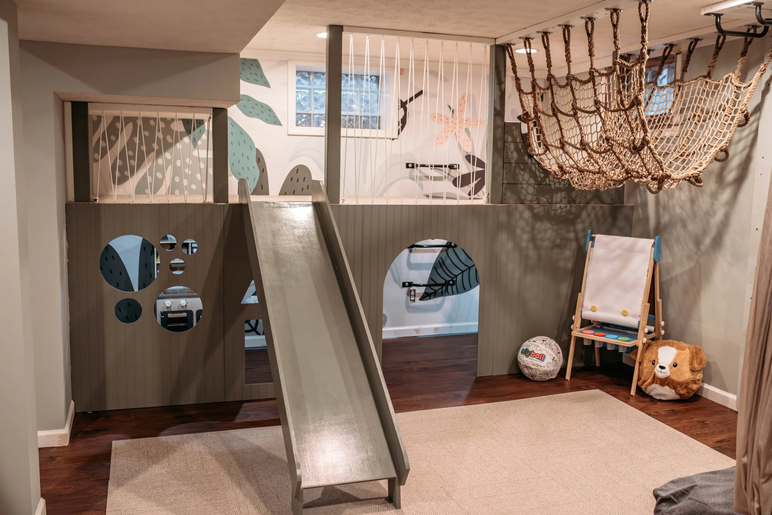 Children's indoor playroom with a slide, climbing structure, and hanging rope net, decorated with playful wall art and toys, including a plush dog and a ball.