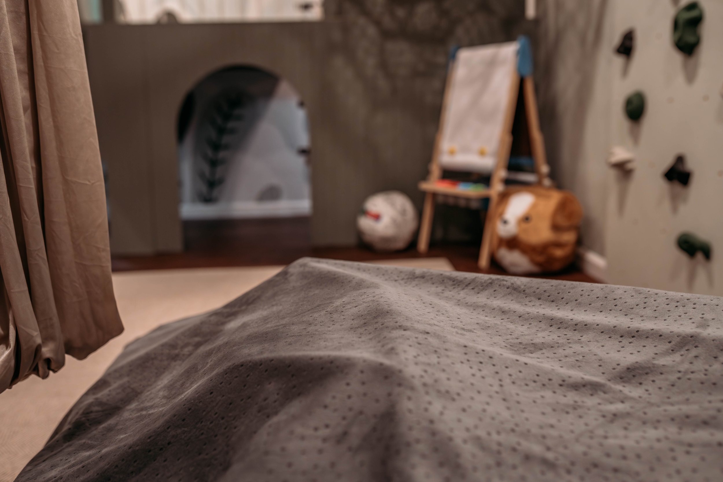 Children's indoor playroom with a crash pad in the foreground, a children's chalkboard easel, plush animal, a climbing wall, and an arched opening in the background.