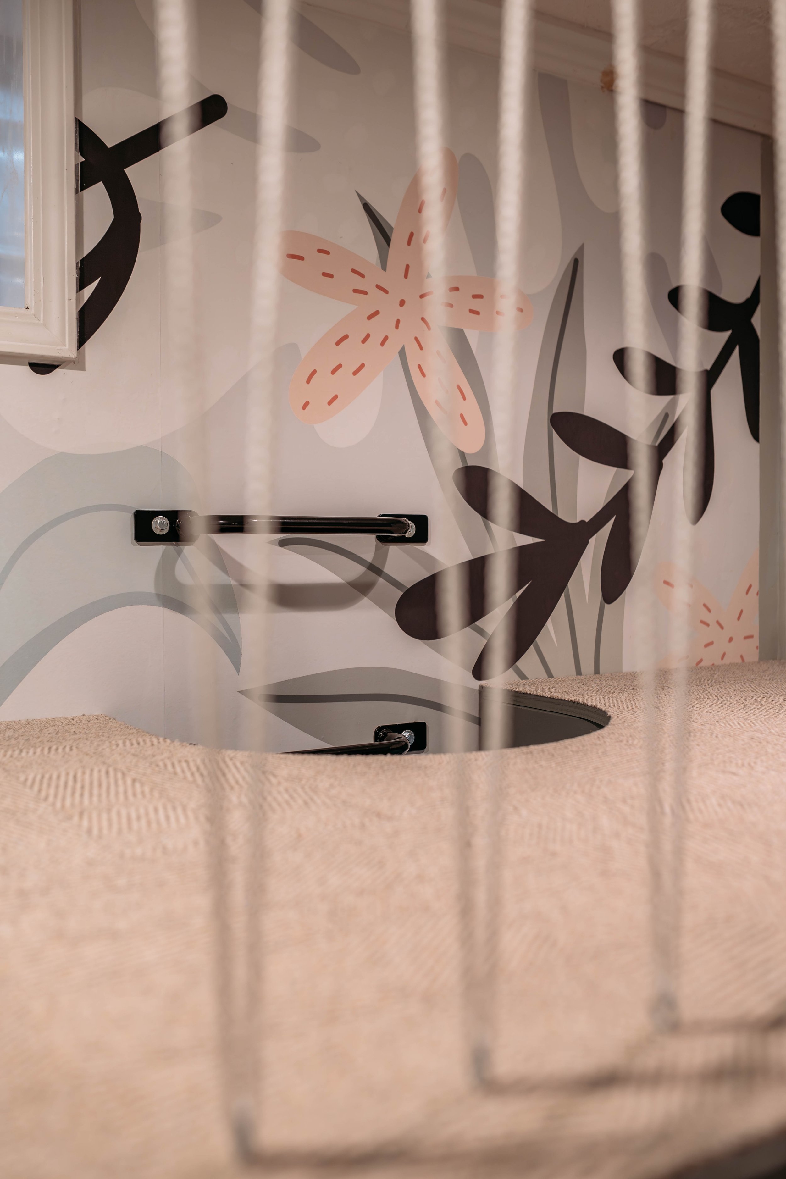 Close-up of a cozy nest with a soft beige carpet, surrounded by vertical white cords; in the background, a wall with a floral mural featuring pink, black, and gray abstract flowers.
