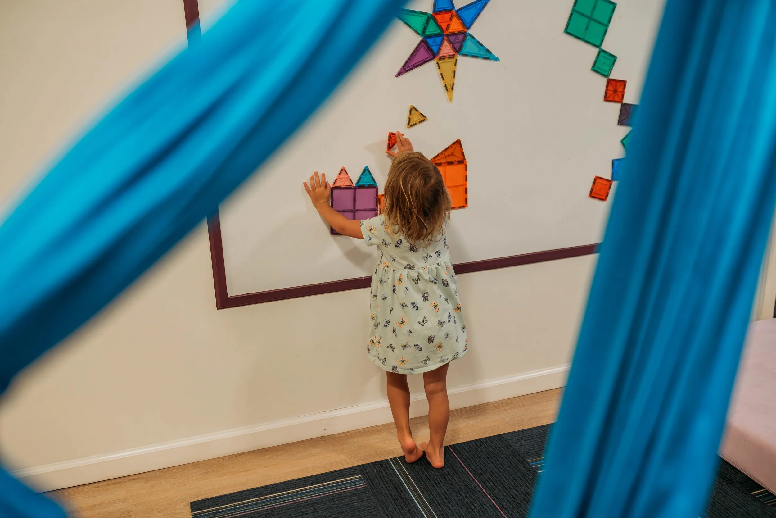 A young girl is playing with colorful magnetic building tiles on a whiteboard, seen through a blue fabric swing.