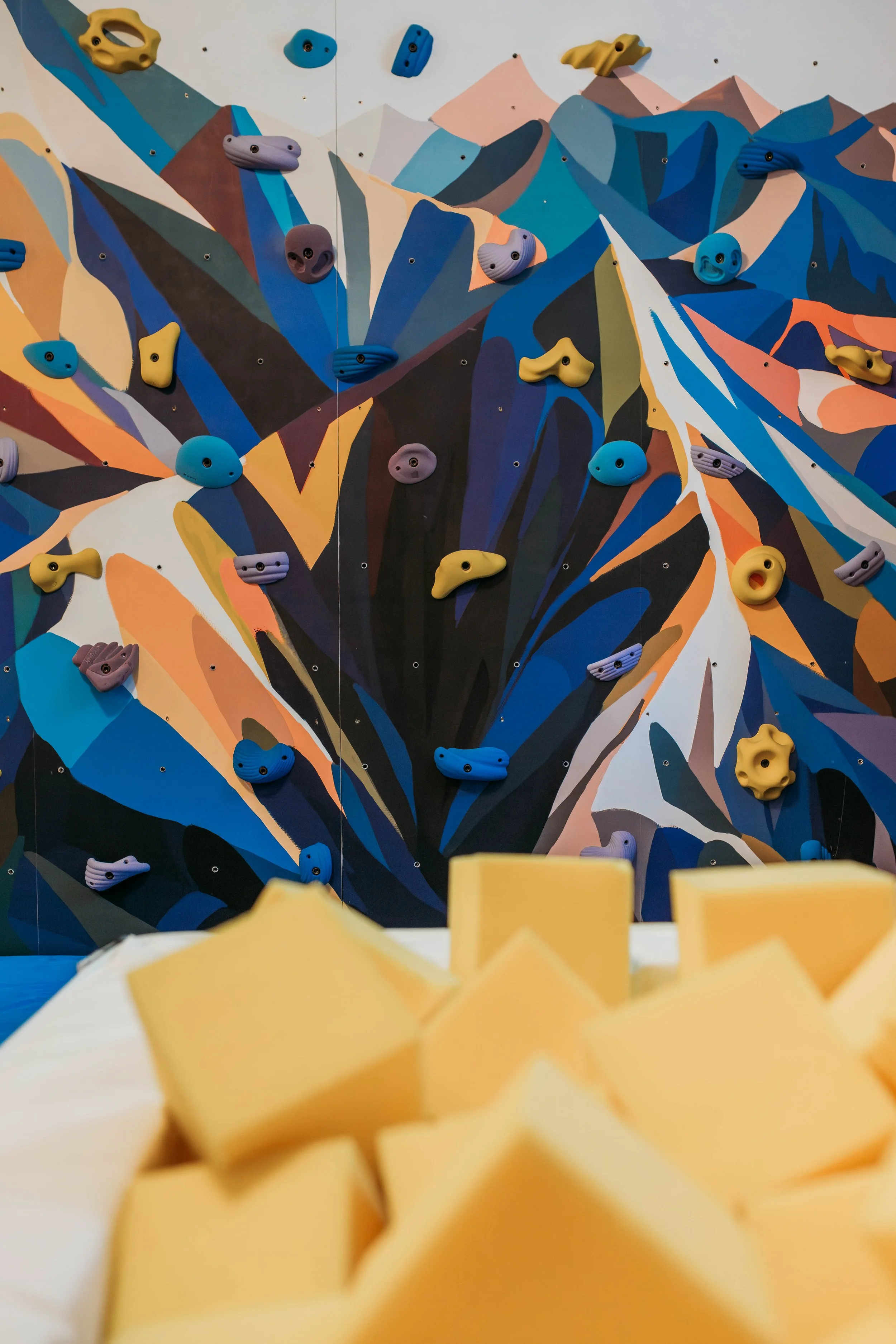 Indoor rock climbing wall with colorful holds and a pile of yellow foam blocks in the foreground.