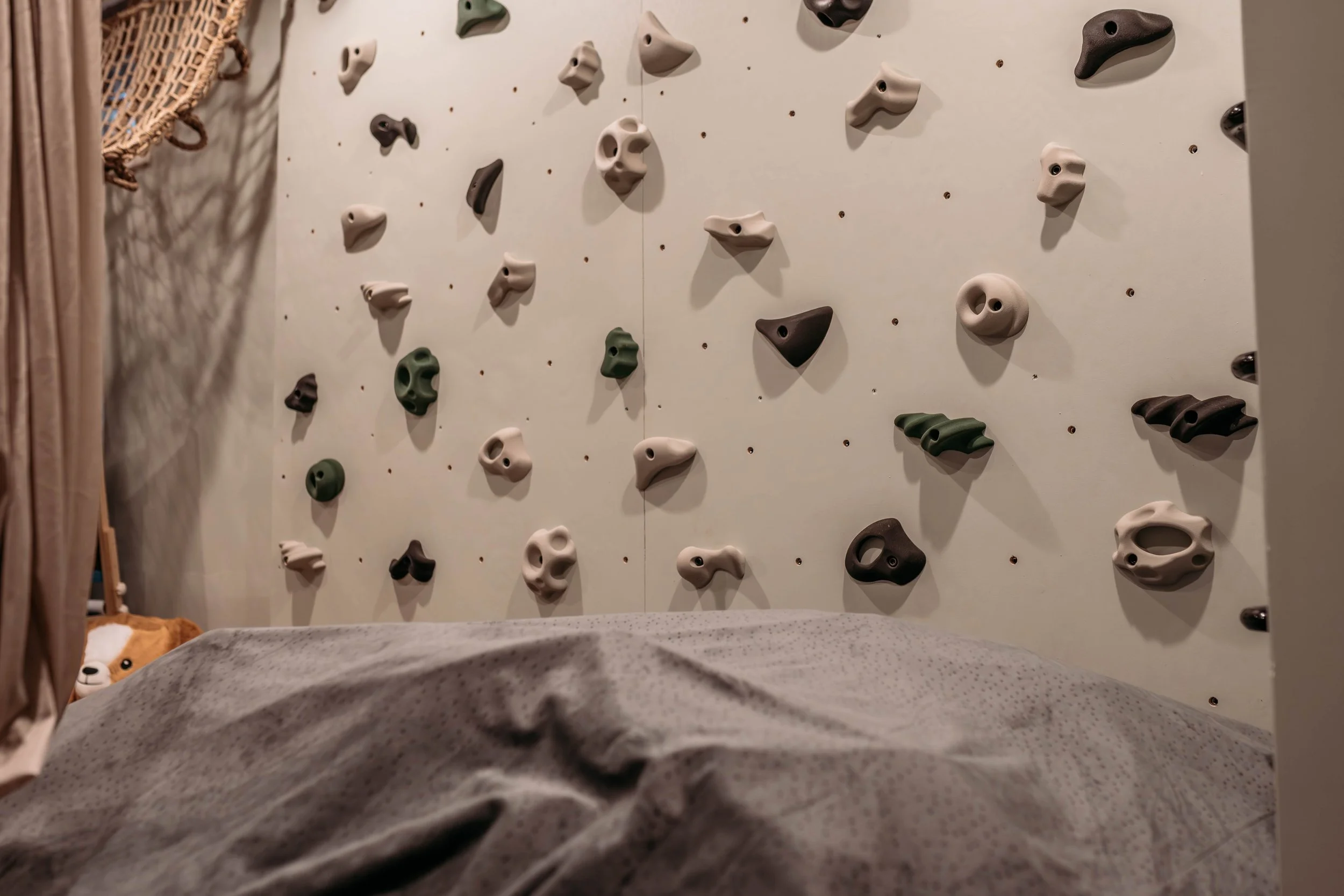 Indoor climbing wall with various colored handholds and footholds, partially obscured crash pad with a stuffed animal peeking out.