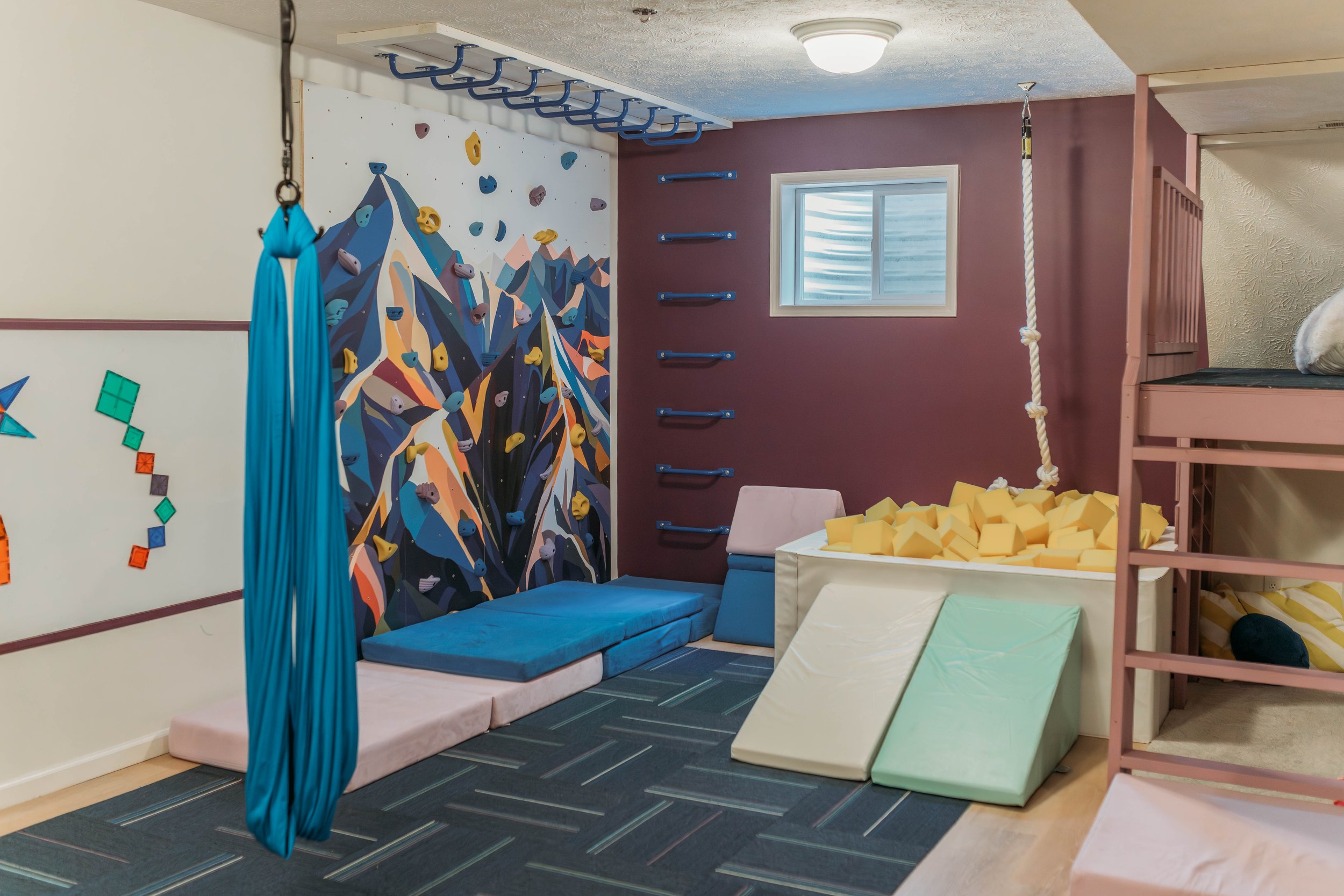 Indoor children's playroom with soft mats, climbing wall, rope, and foam blocks.