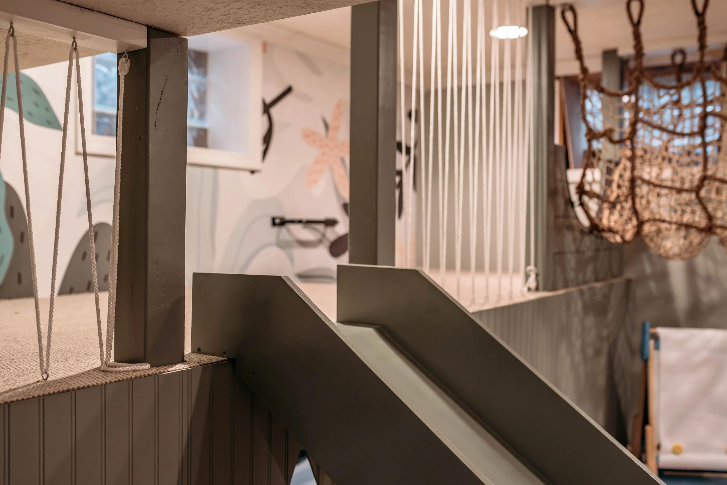 Indoor play area with a wood slide, rope climbing structures, and painted wall with floral and abstract designs.
