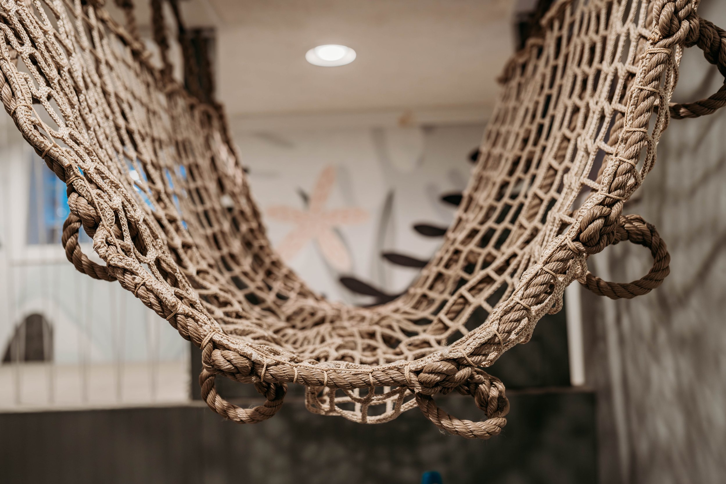 Close-up view of a woven rope hammock hanging indoors, with a blurred wall in the background.