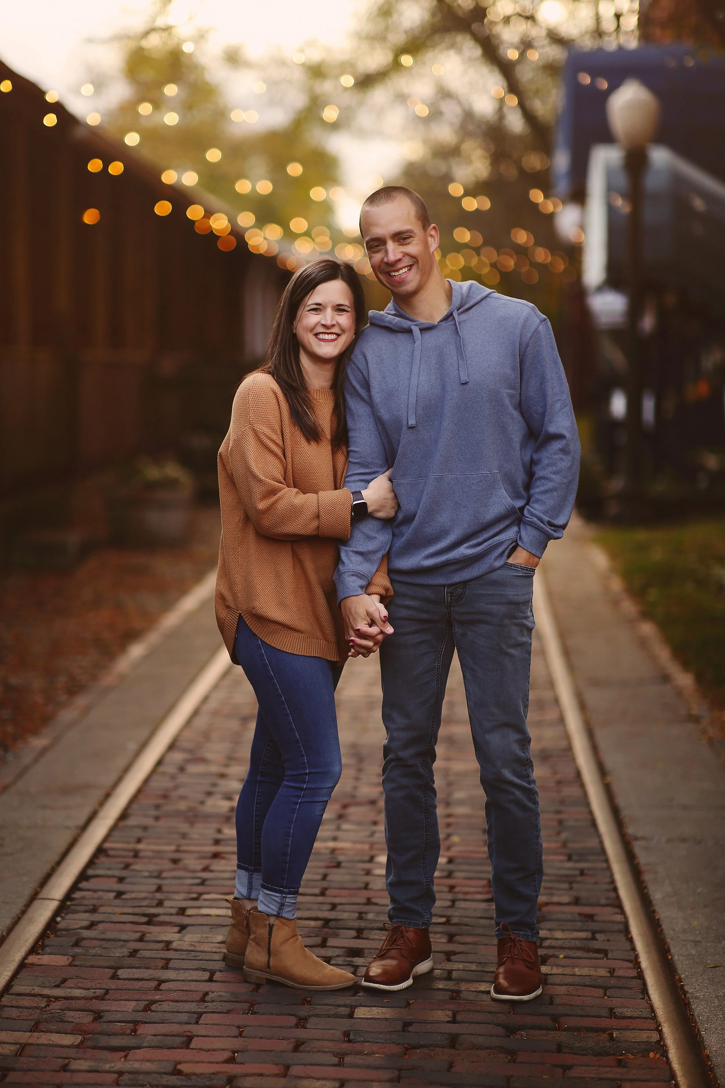 A smiling couple holding hands on a brick pathway outdoors at dusk, with string lights hanging above and trees in the background.