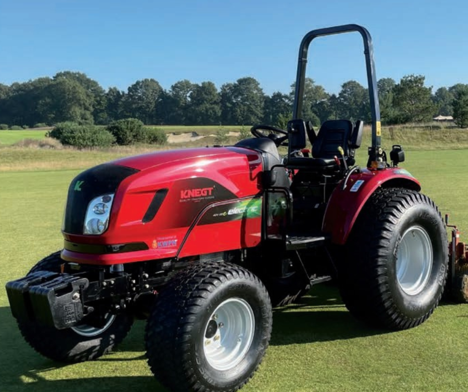 Red tractor on a grassy field with trees and clear blue sky in the background.